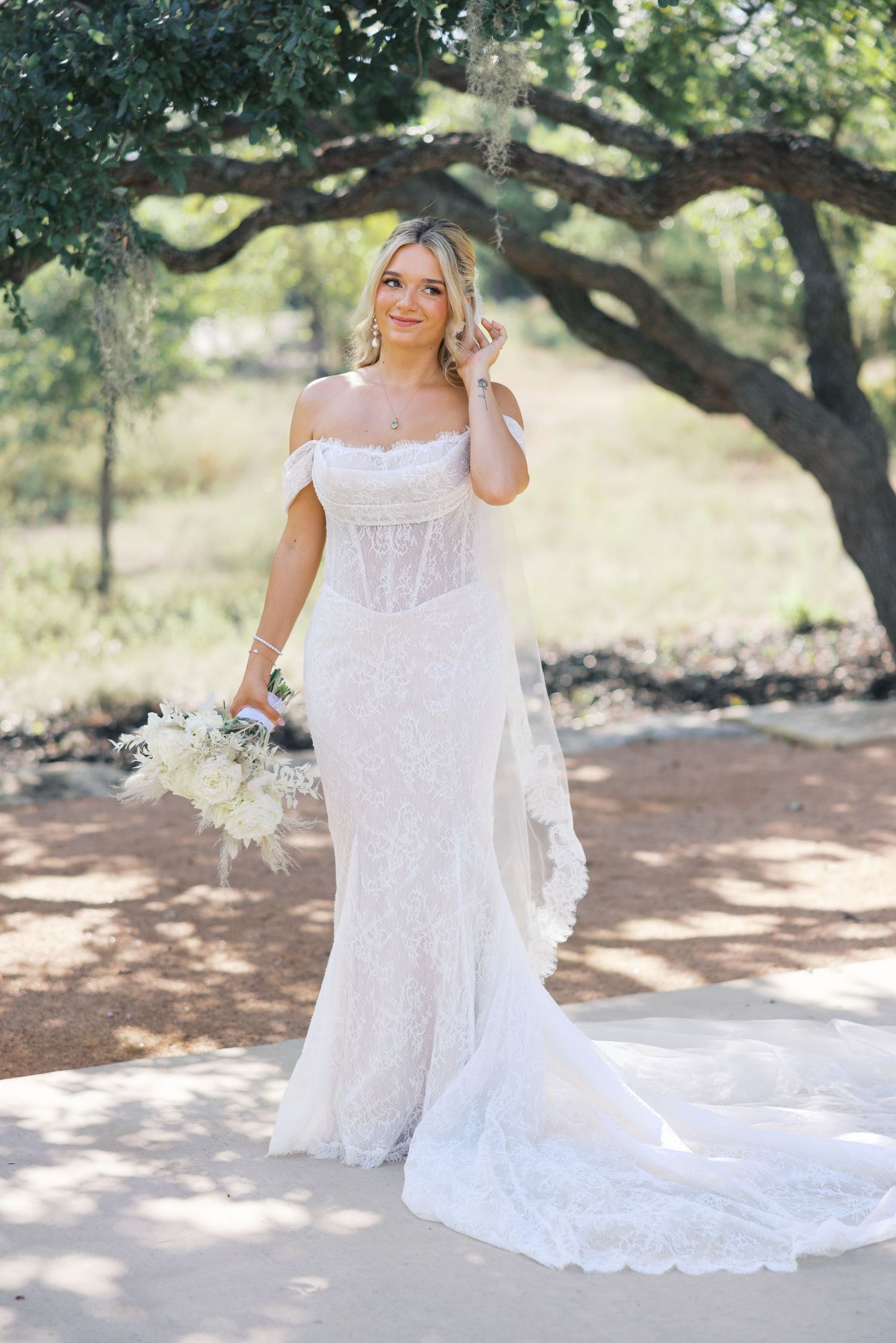 Bride in corseted lace gown holds white bouquet while posing under oak tree branches.