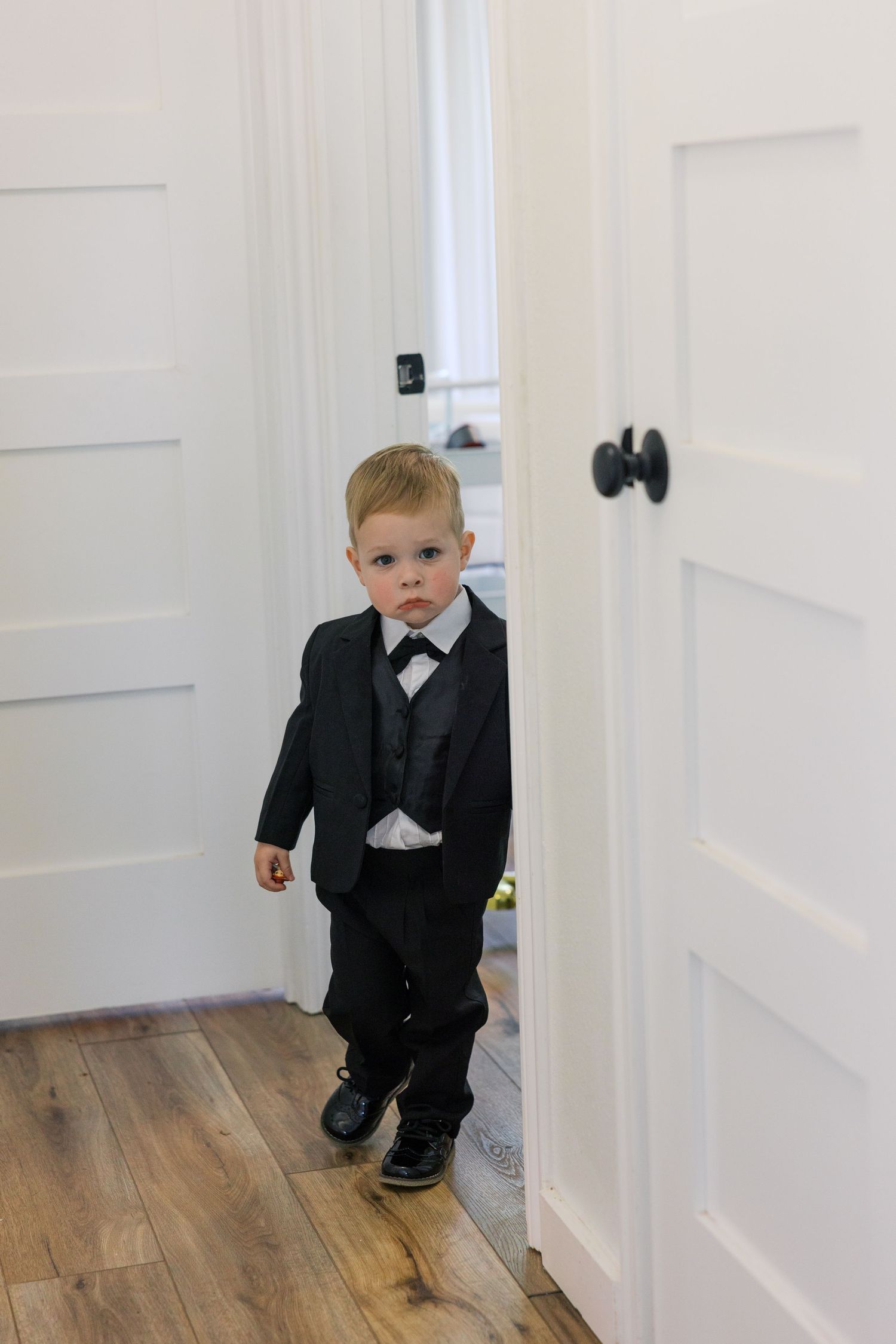 Young boy in black tuxedo suit standing in doorway on hardwood floor, dressed for formal occasion.