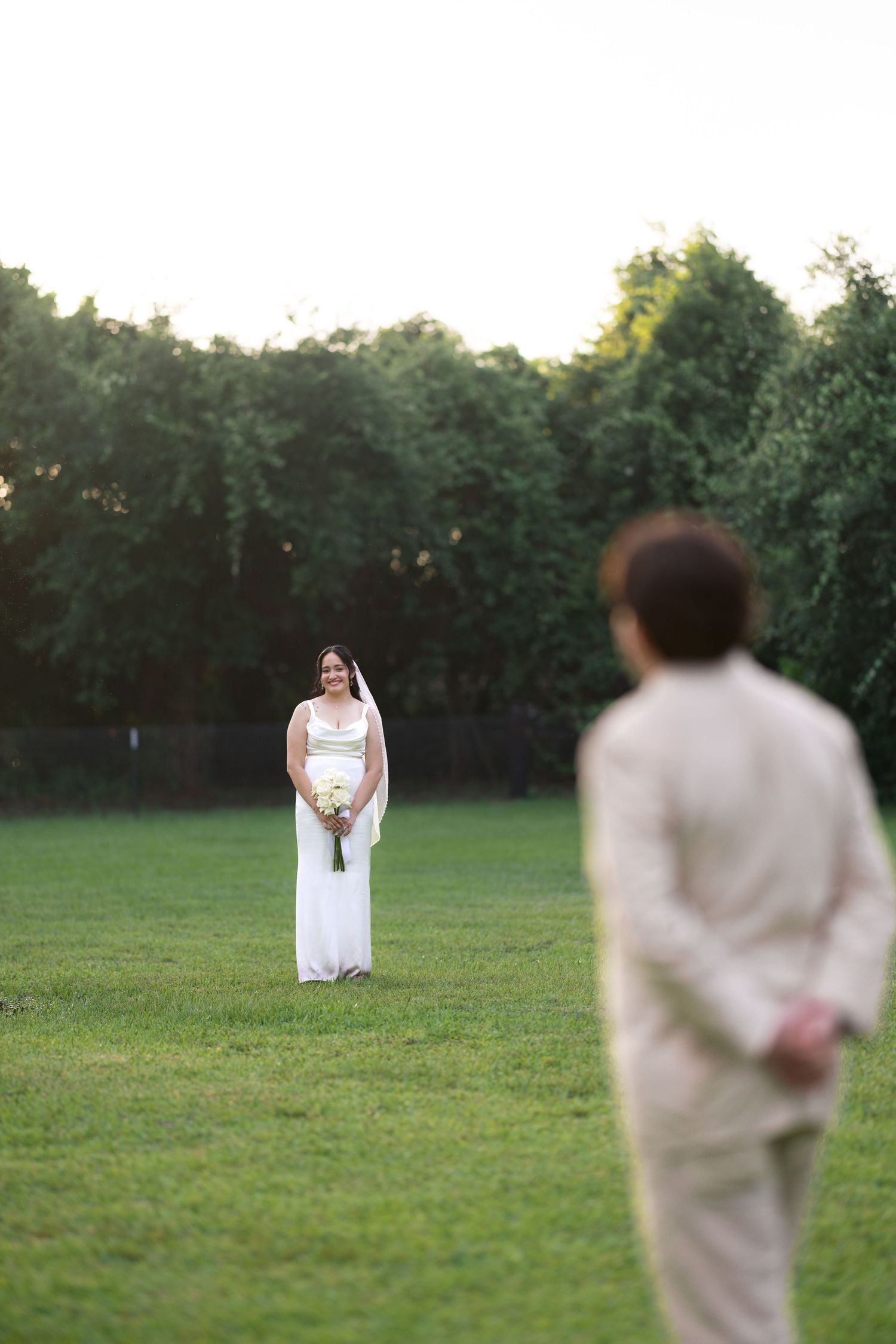 Bride in white dress holding bouquet faces groom in beige suit during first look on green lawn.