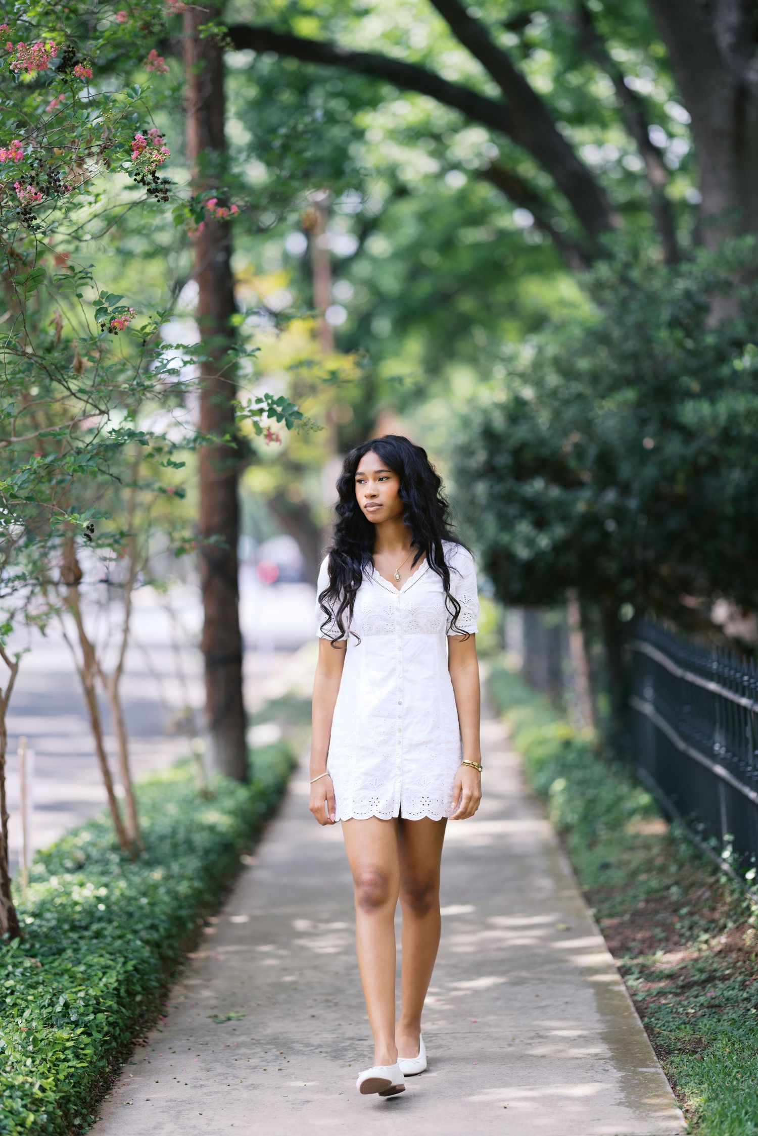 Young Black woman in white mini dress walks along tree-lined urban sidewalk path.