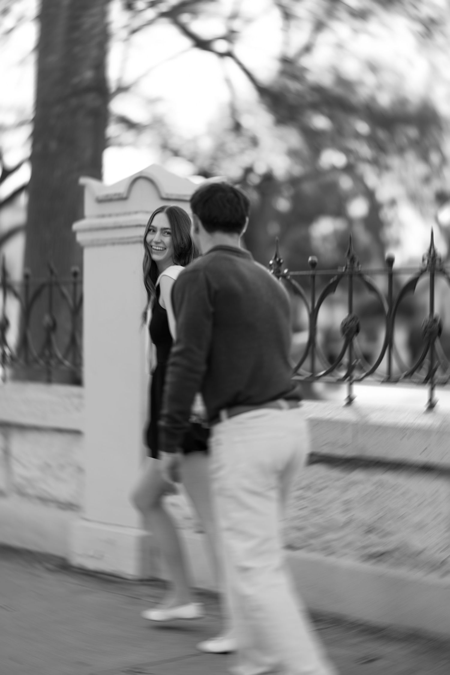 Black and white photo of a smiling young woman walking with a man along an iron fence outdoors.