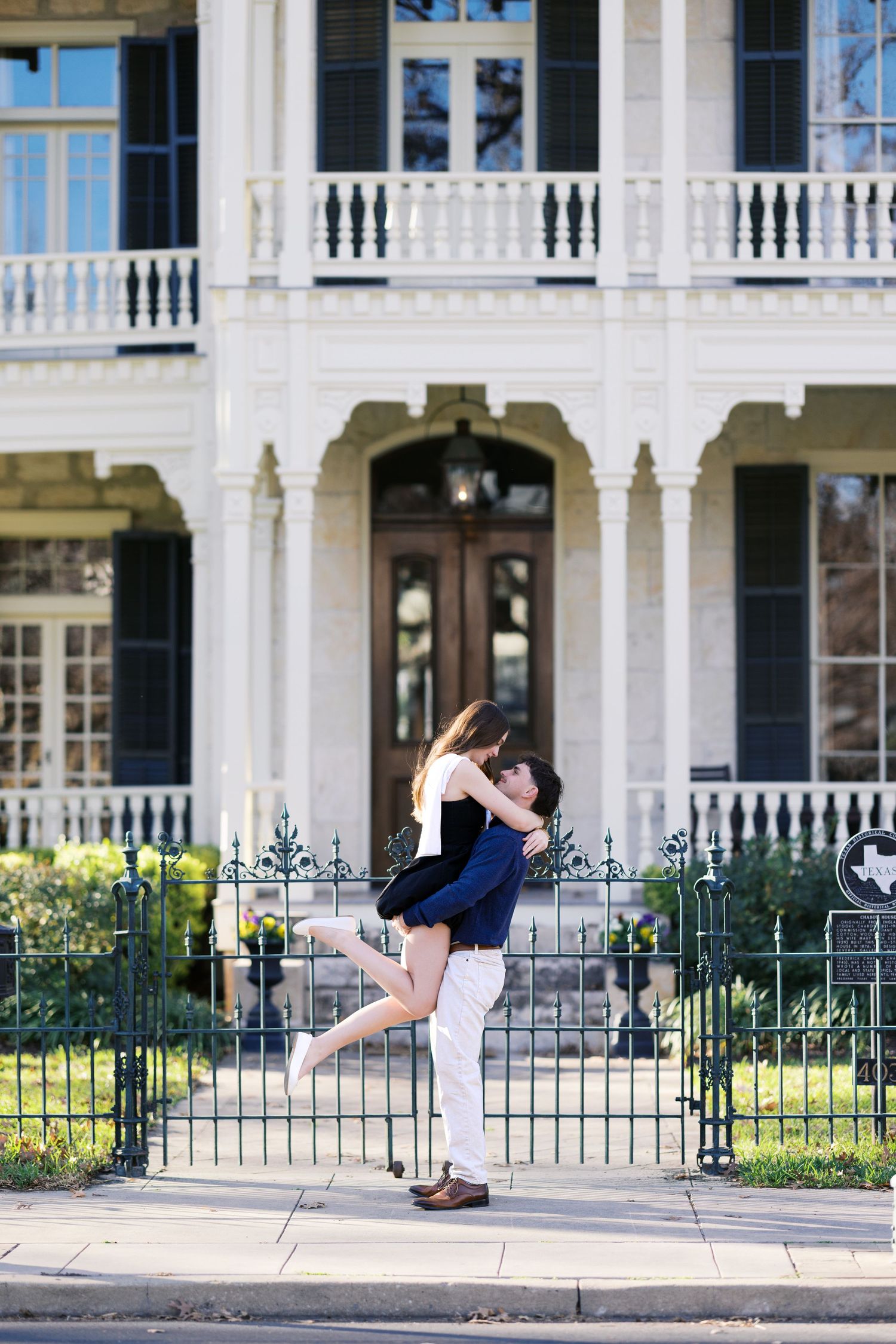 Couple embracing in front of a white Southern mansion with ornate balconies and iron fence.