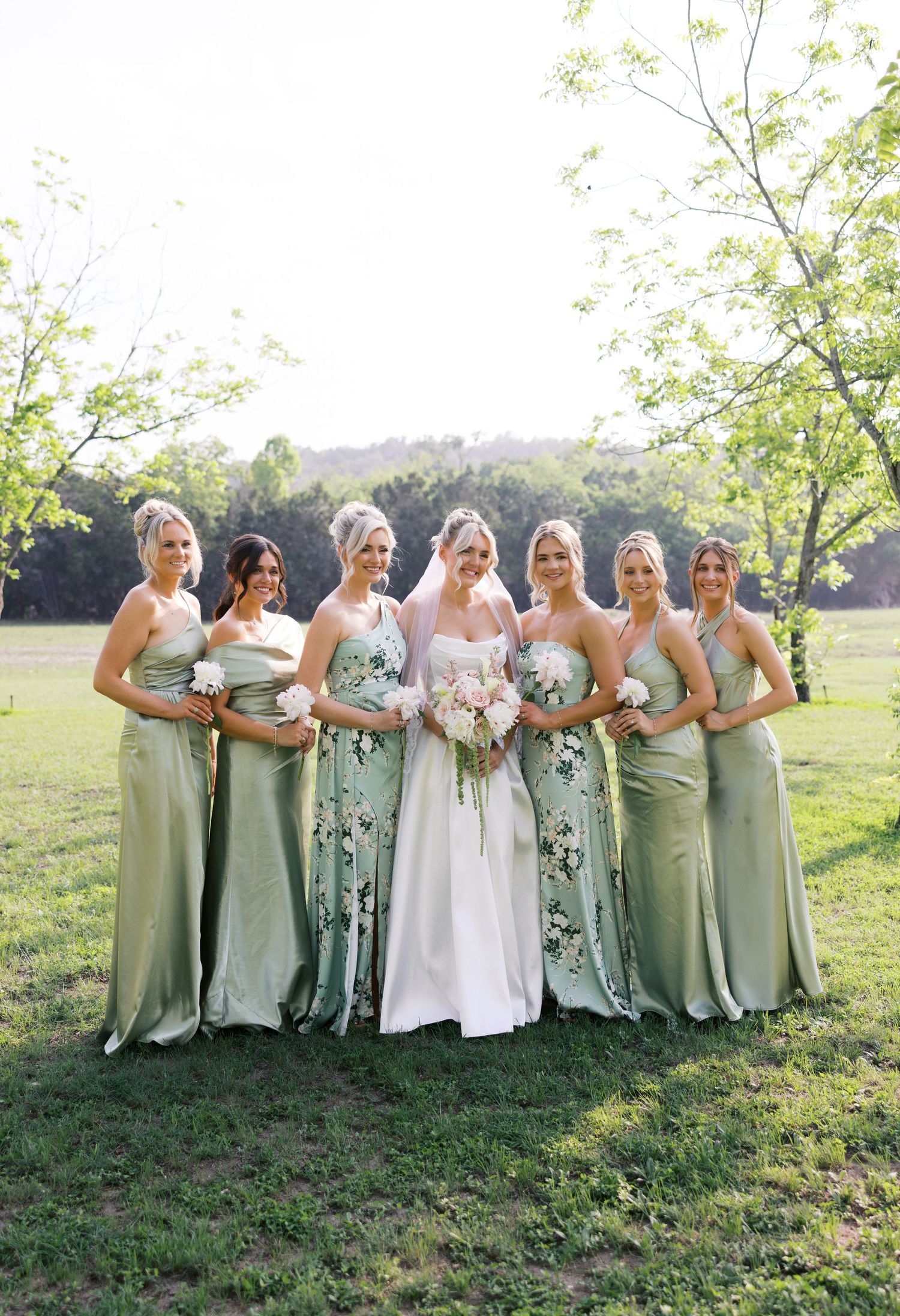 Bride in floral white gown poses with five bridesmaids in sage green satin dresses outdoors on green lawn.