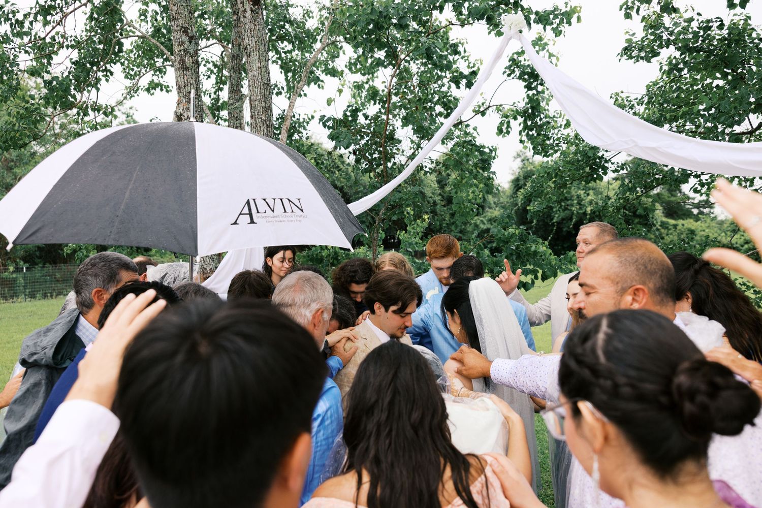 Outdoor wedding ceremony crowd gathered under black and white Alvin umbrella amid lush green trees.