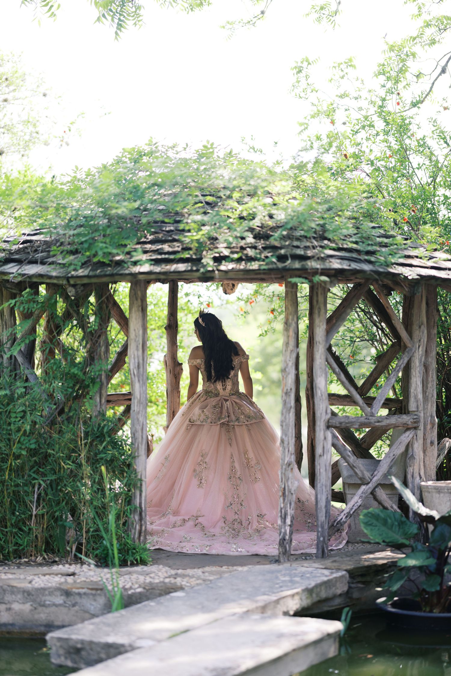 Woman in pink ballgown standing in rustic wooden gazebo surrounded by lush greenery.
