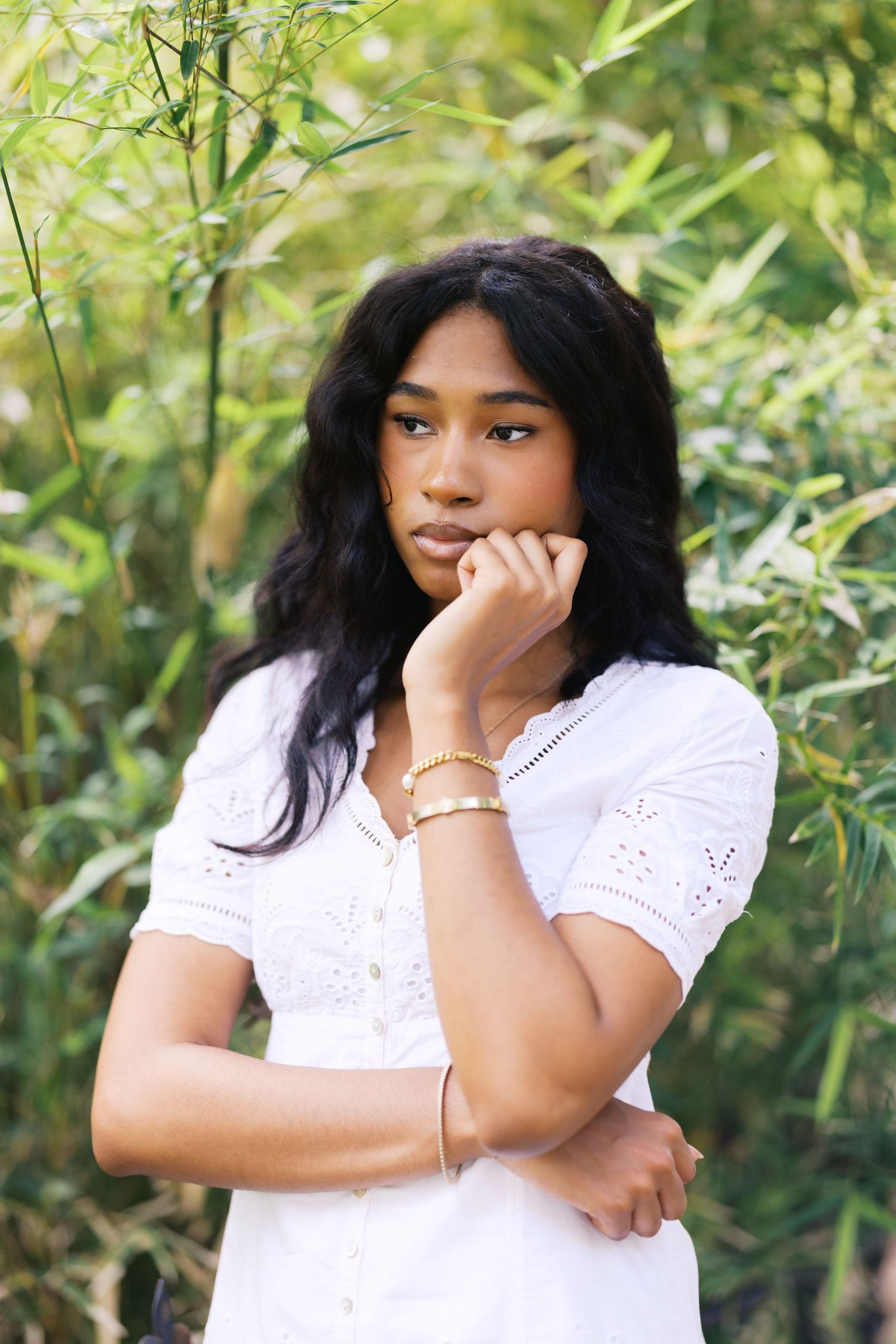 Young woman in white eyelet dress posing thoughtfully with hand on chin among bamboo plants.