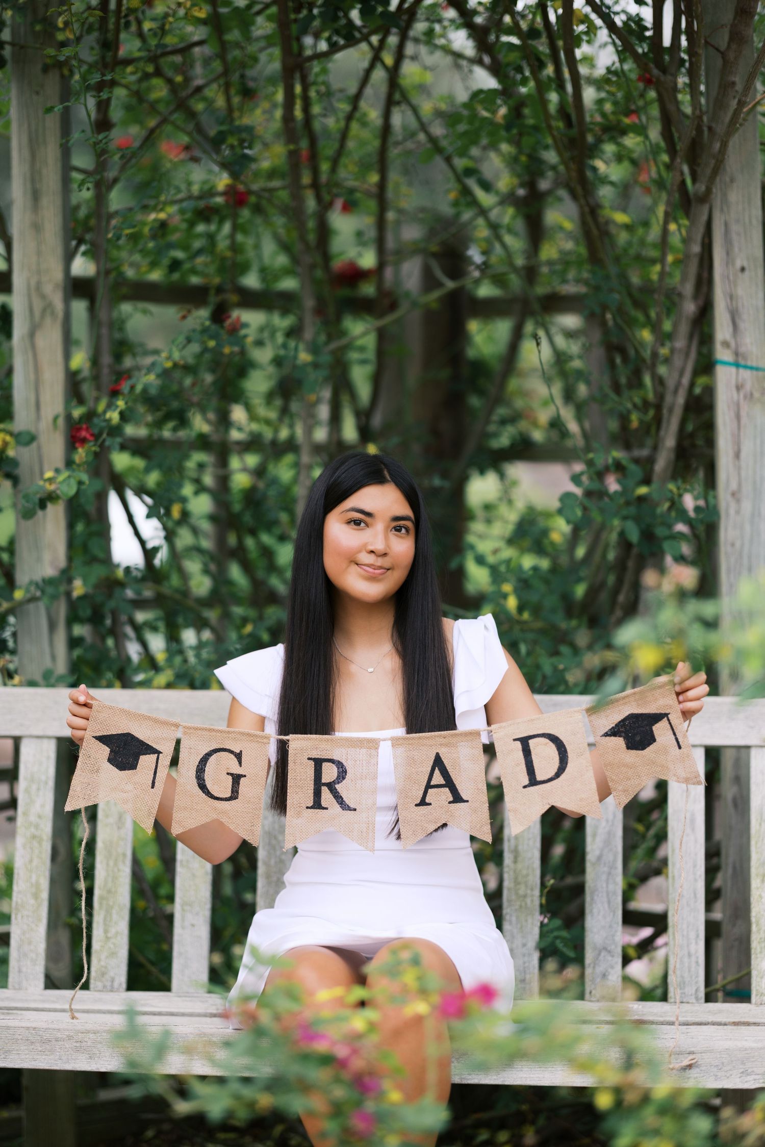 Young woman in white dress holding a burlap GRAD banner, sitting on a wooden bench surrounded by lush greenery.