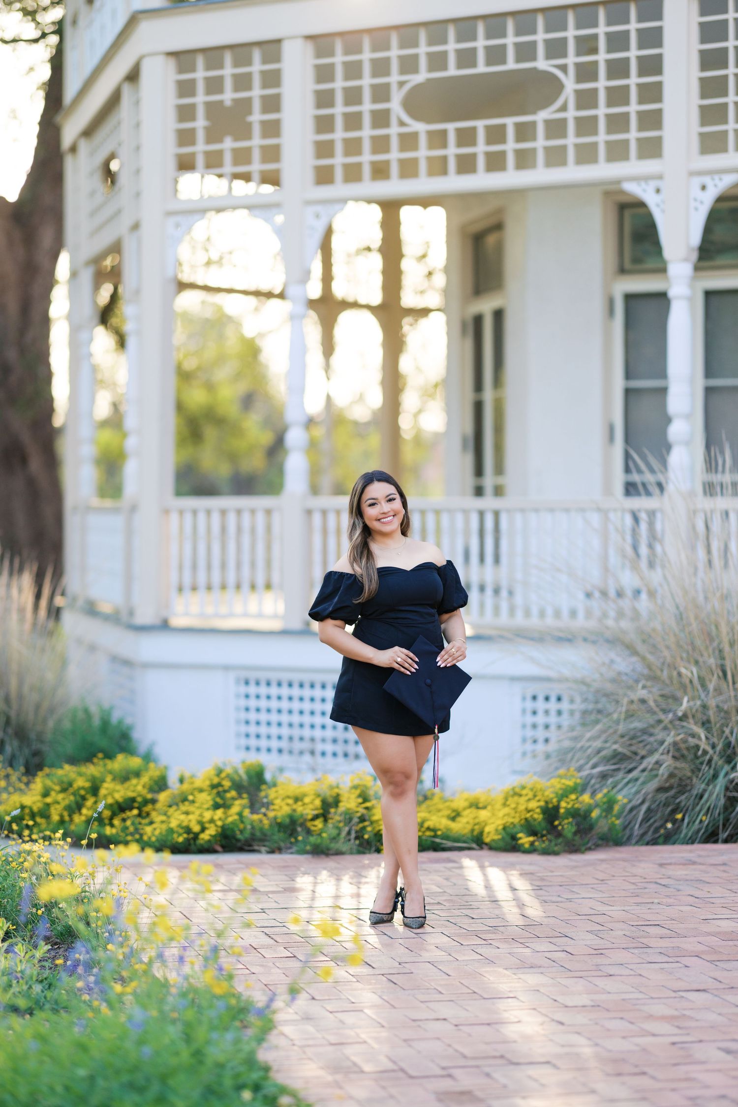 Young woman in black off-shoulder mini dress poses on brick path by white Victorian gazebo surrounded by yellow flowers.