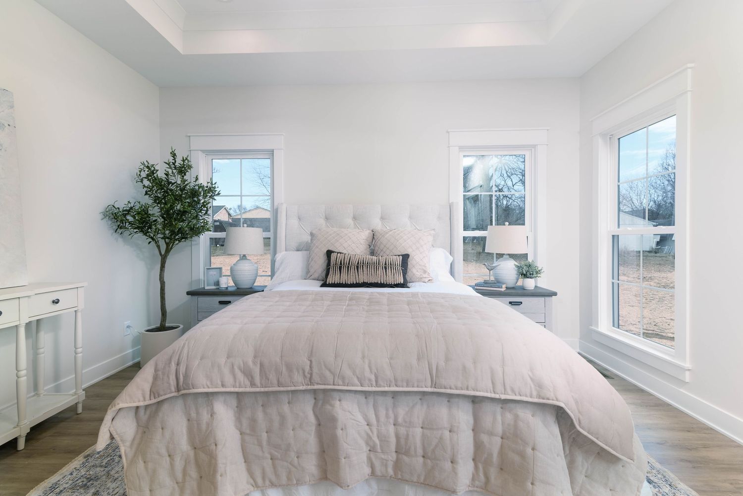 Bright master bedroom with tray ceiling, upholstered headboard, twin windows, and hardwood floors.
