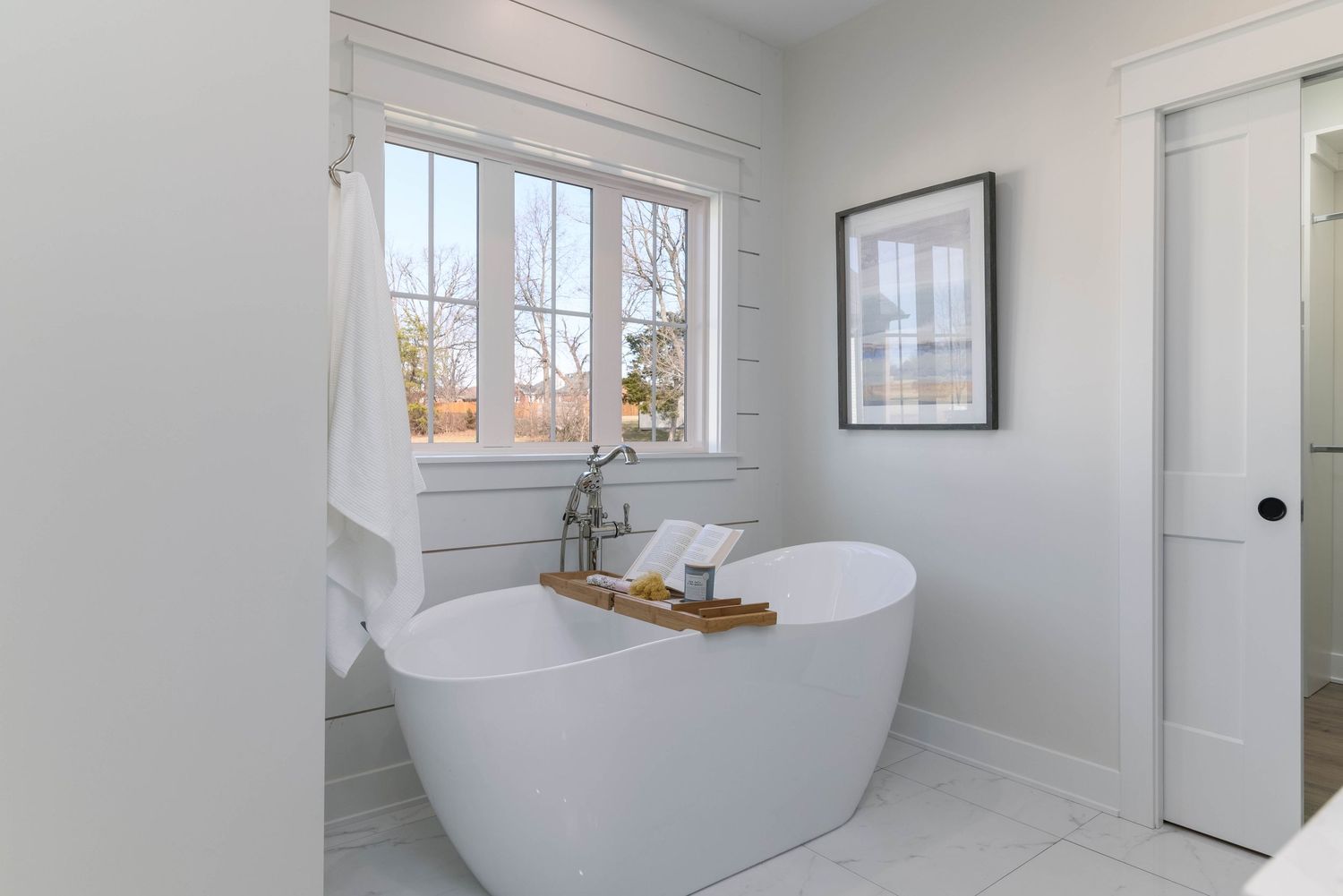 Bright white bathroom featuring a freestanding oval soaking tub beneath a large window with natural light.