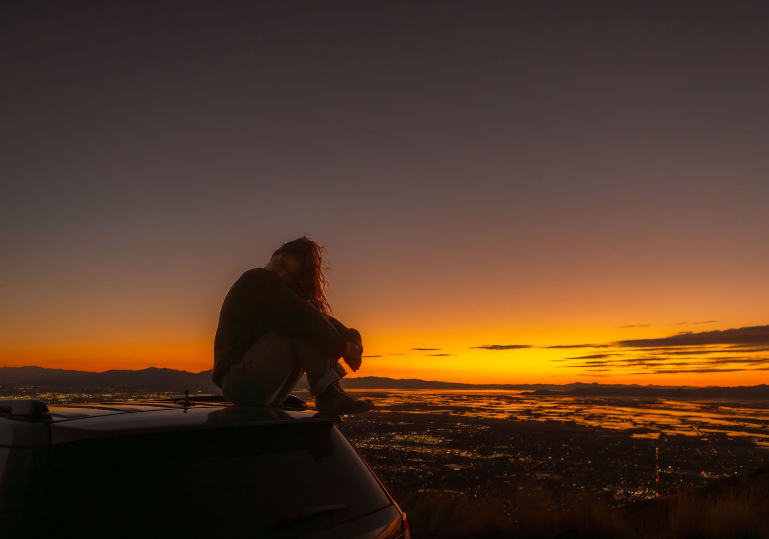 girl poses on top of car during moody sunset overlooking city and mountains in utah