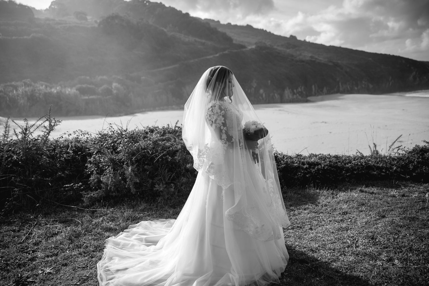 A bride in a flowing white wedding dress stands in a scenic outdoor field against a dramatic cloudy sky.