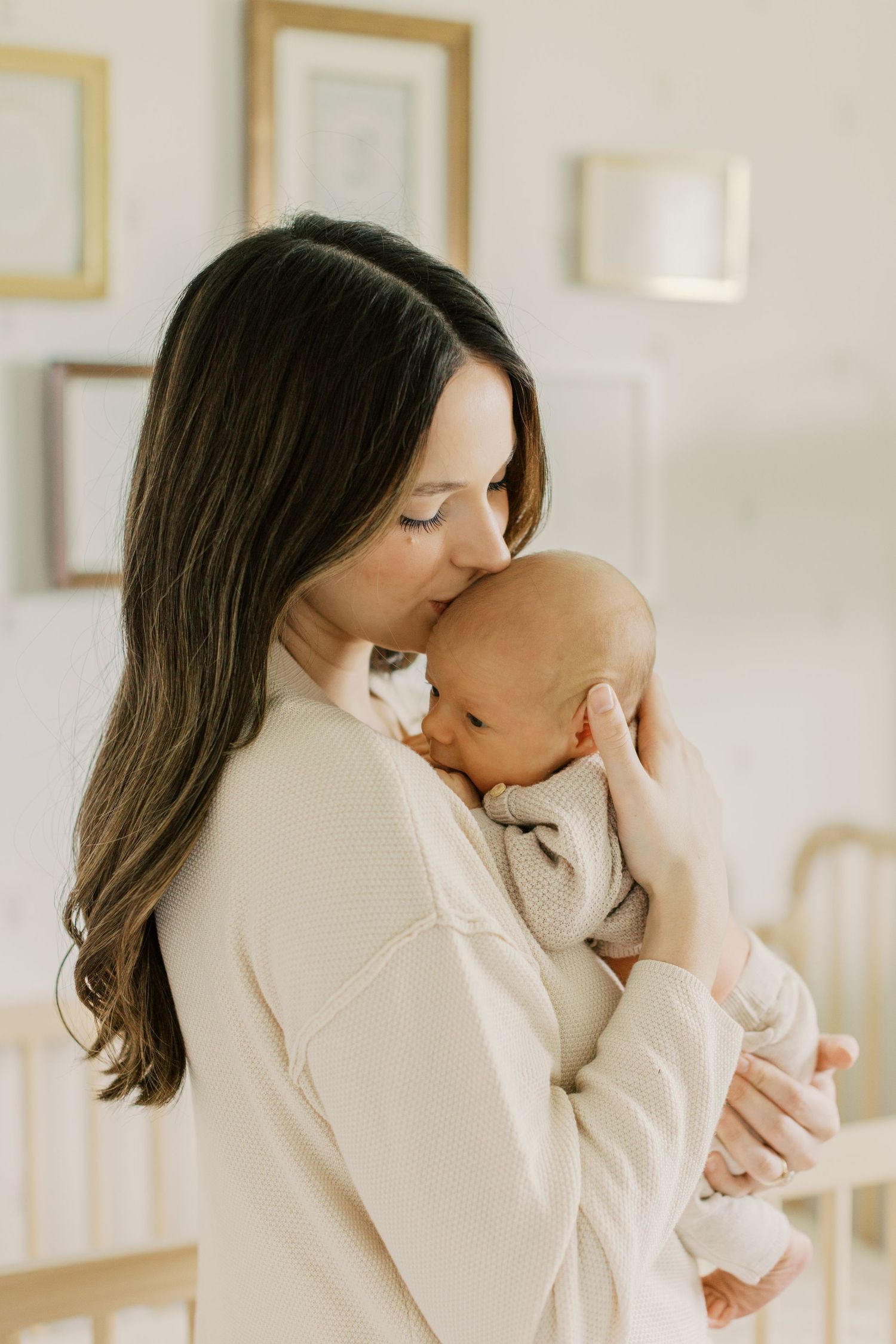 A mother tenderly kisses her newborn baby while cradling them close in a softly lit nursery room.