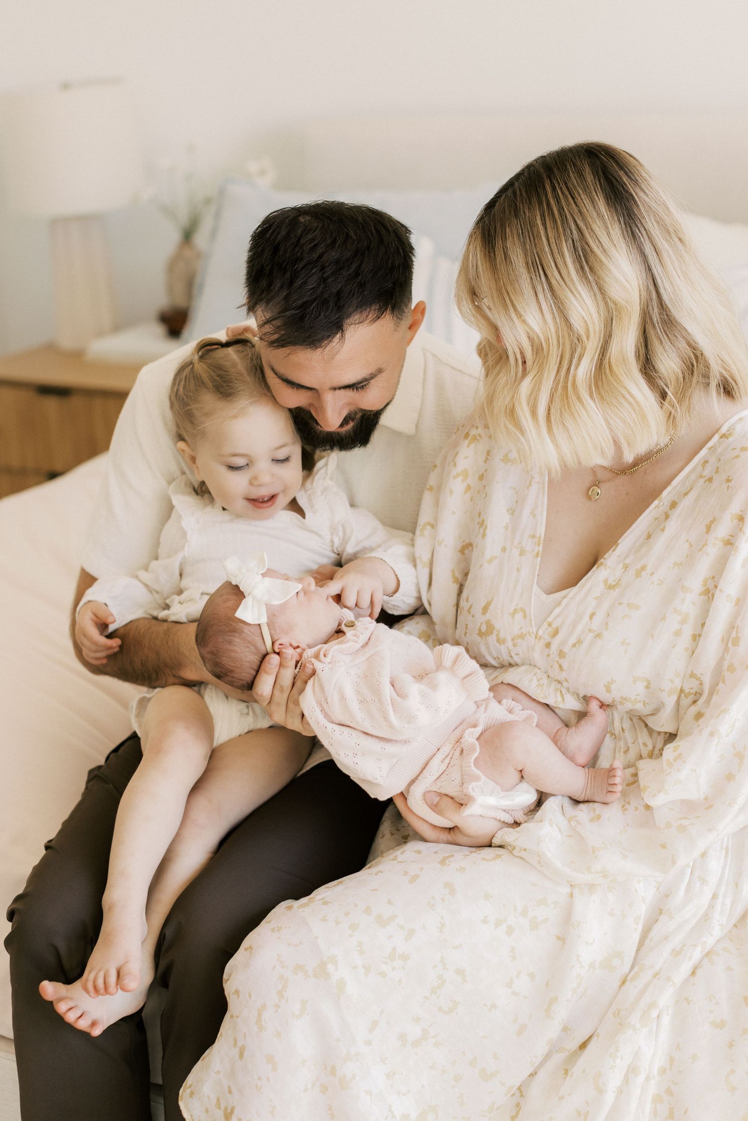 A young family sits together on a bed, parents smiling at their toddler holding a newborn baby in pink.