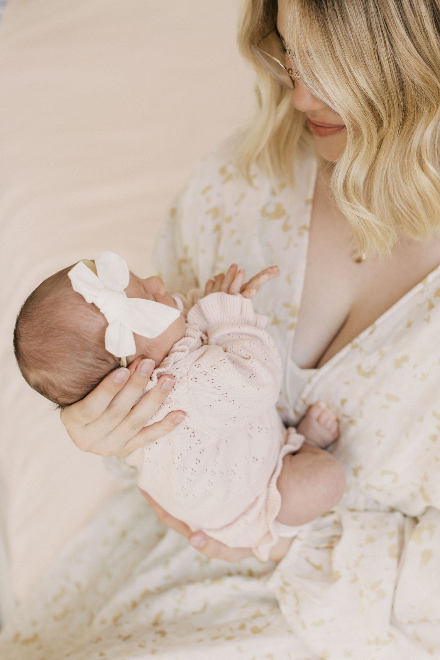 A blonde mother cradles her newborn baby girl dressed in pink with a white bow headband.