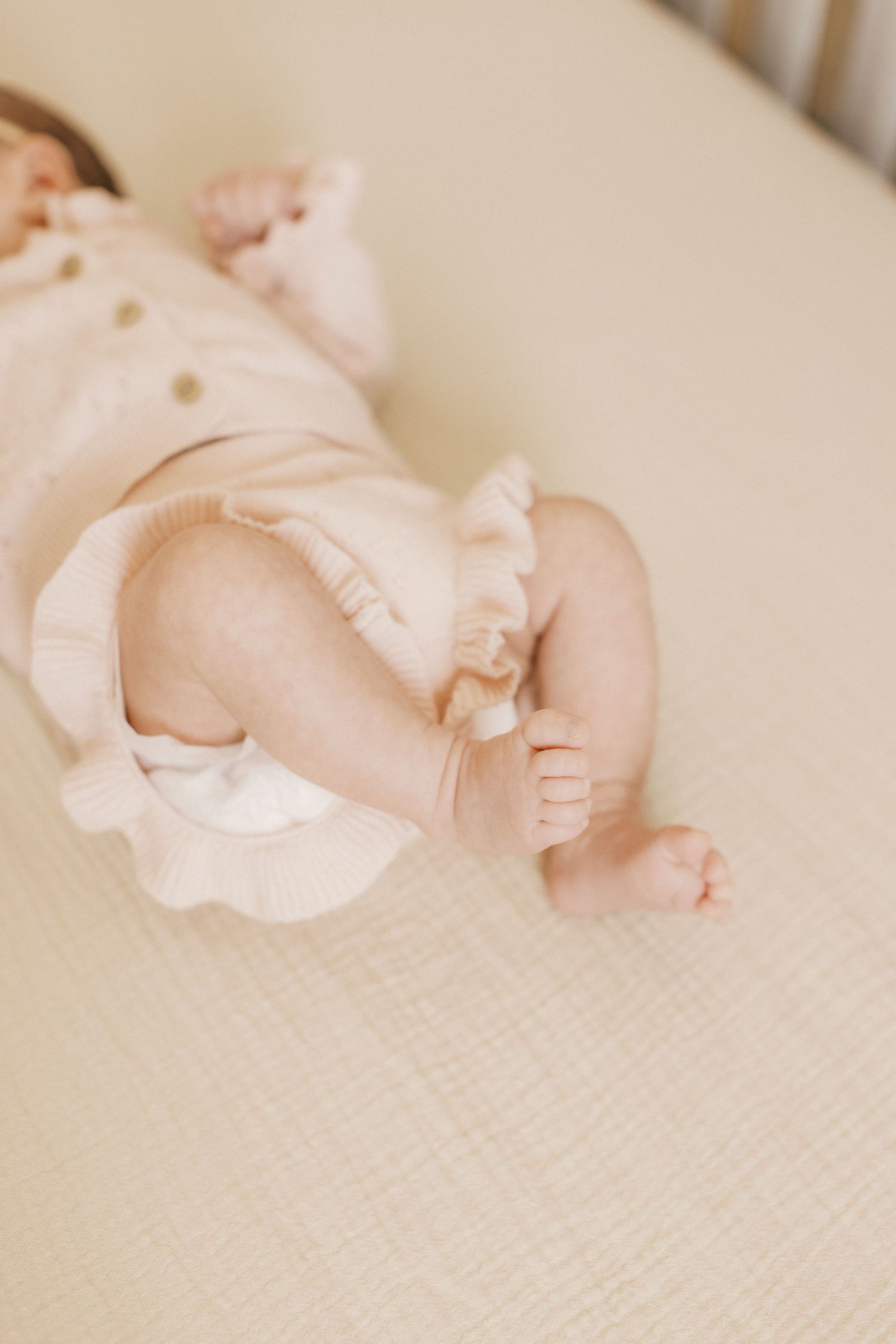 Newborn baby in pink ruffled outfit lying on a cream muslin crib sheet, showing chubby legs and tiny toes.