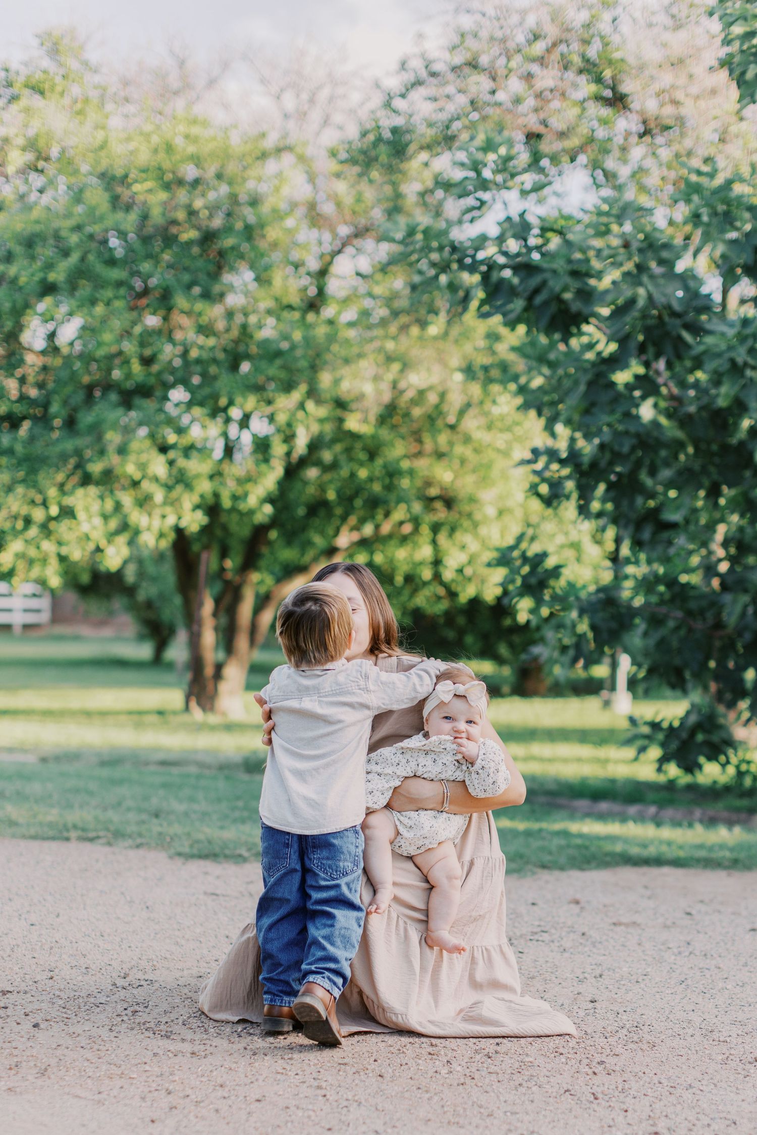 A child hugs a woman sitting on a path holding a baby, surrounded by lush green trees in a park.
