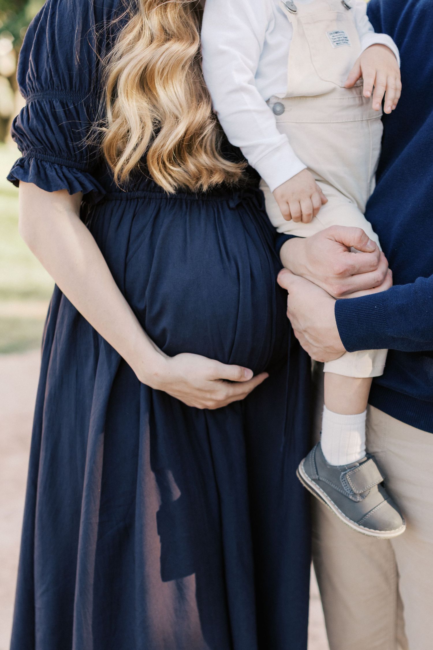 Pregnant woman in navy dress cradling baby bump beside partner holding toddler in white outfit outdoors.