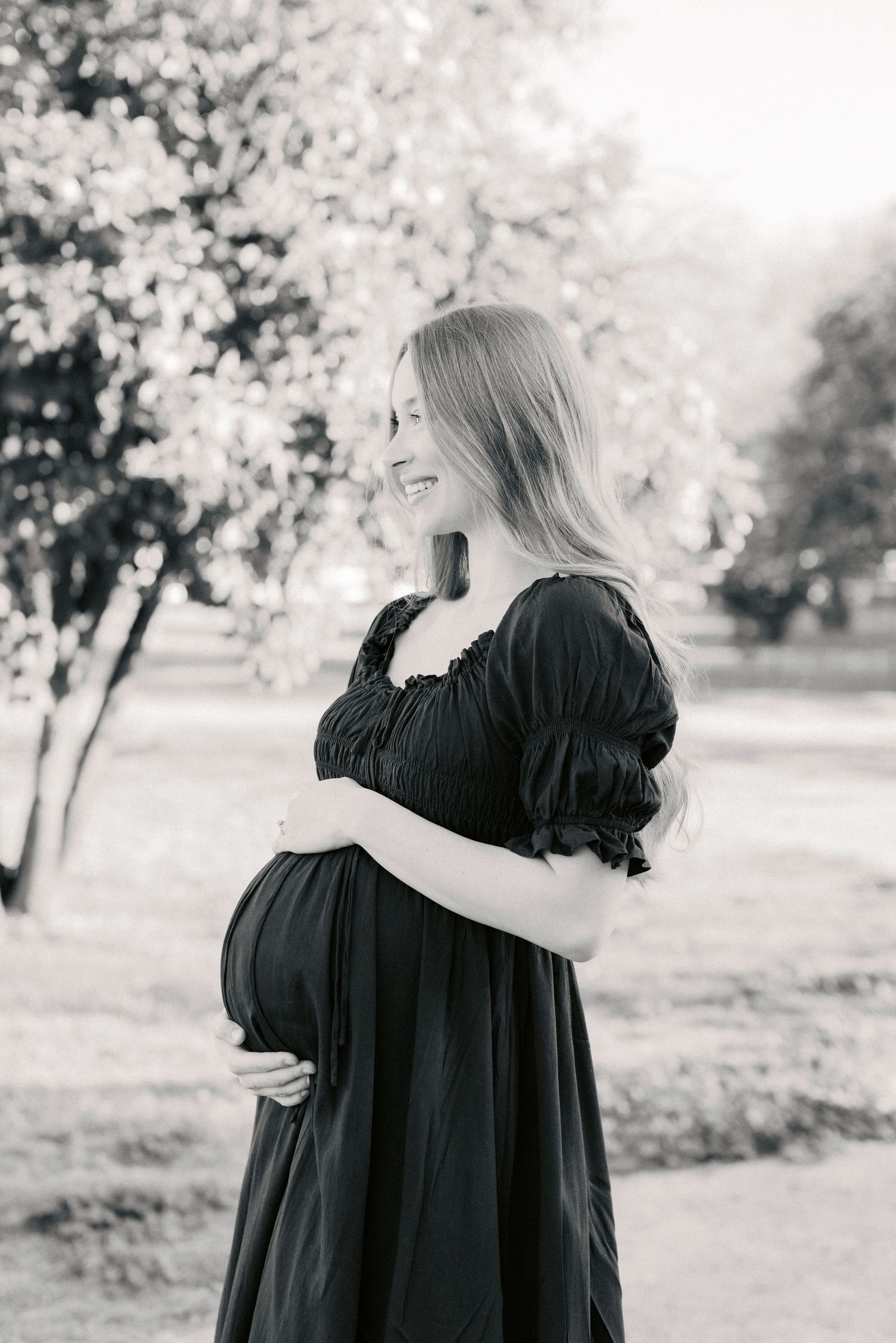 Black and white photo of a smiling pregnant woman in a dark dress cradling her belly outdoors in a sunny park.