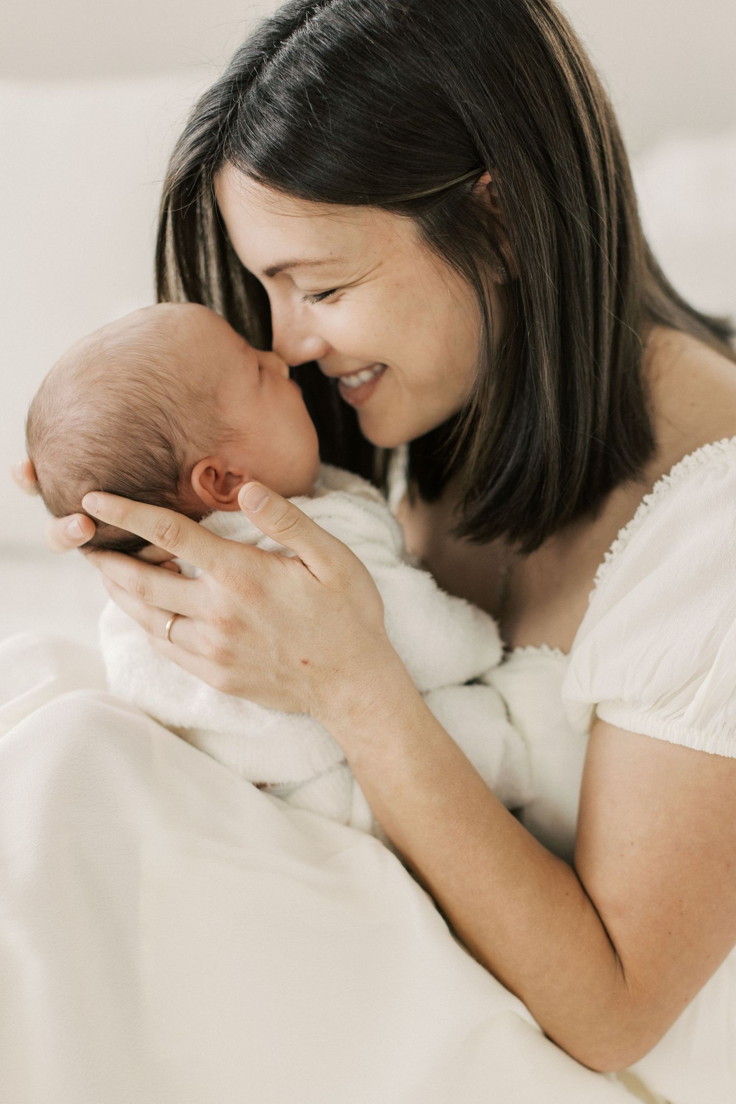 A smiling mother in a white dress tenderly holds her newborn baby close, touching noses in a bright, soft setting.