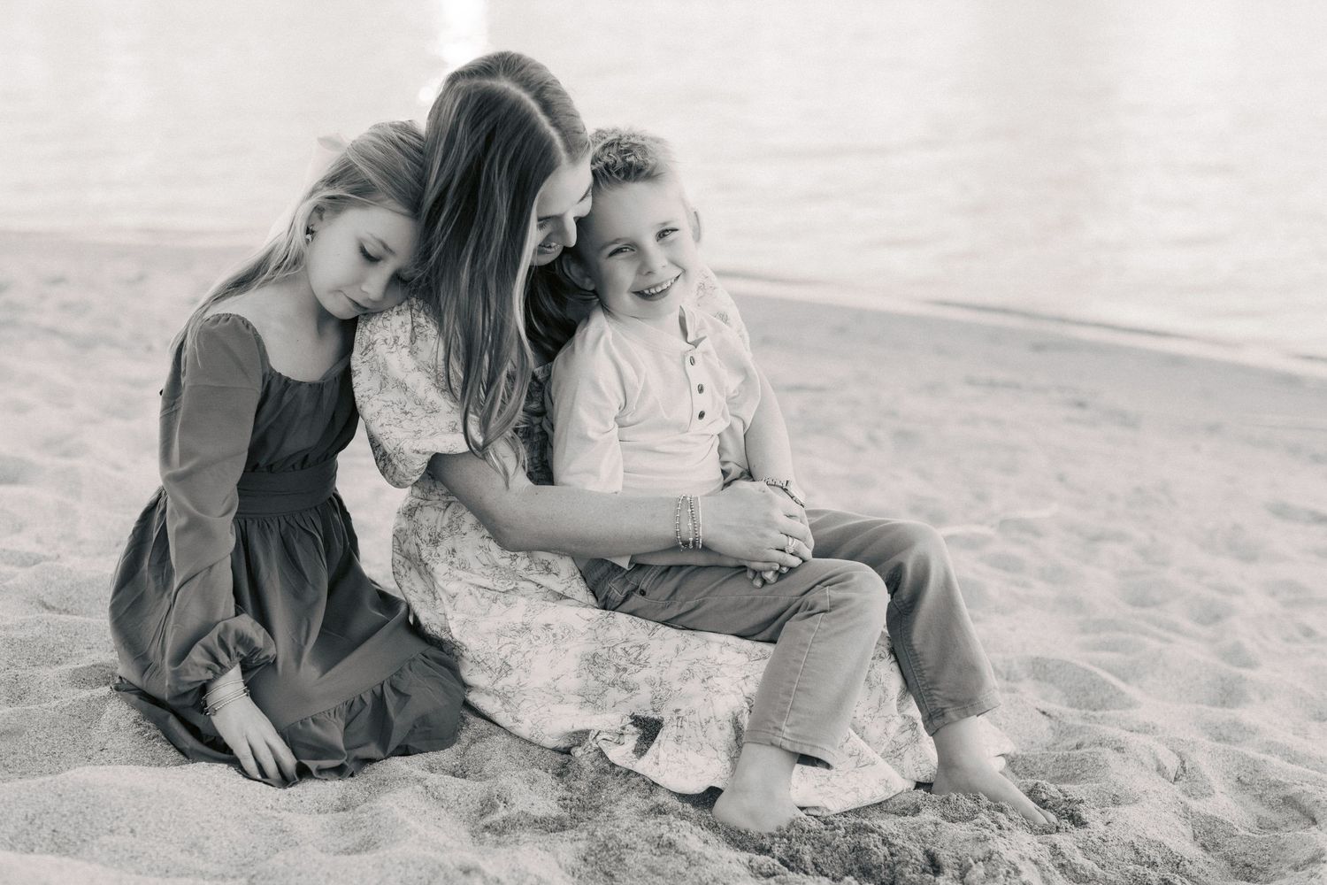 Black and white photo of a mother and two children sitting together on a sandy beach, smiling warmly.