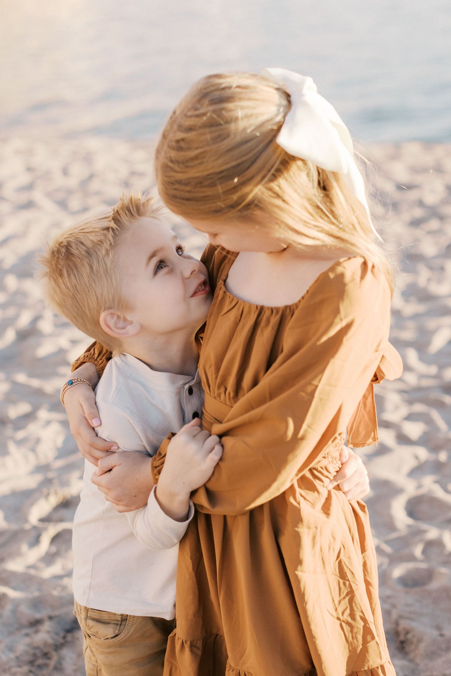 Two young siblings share a sweet embrace on a sandy beach, with a girl in a rust dress hugging a boy in warm sunlight.