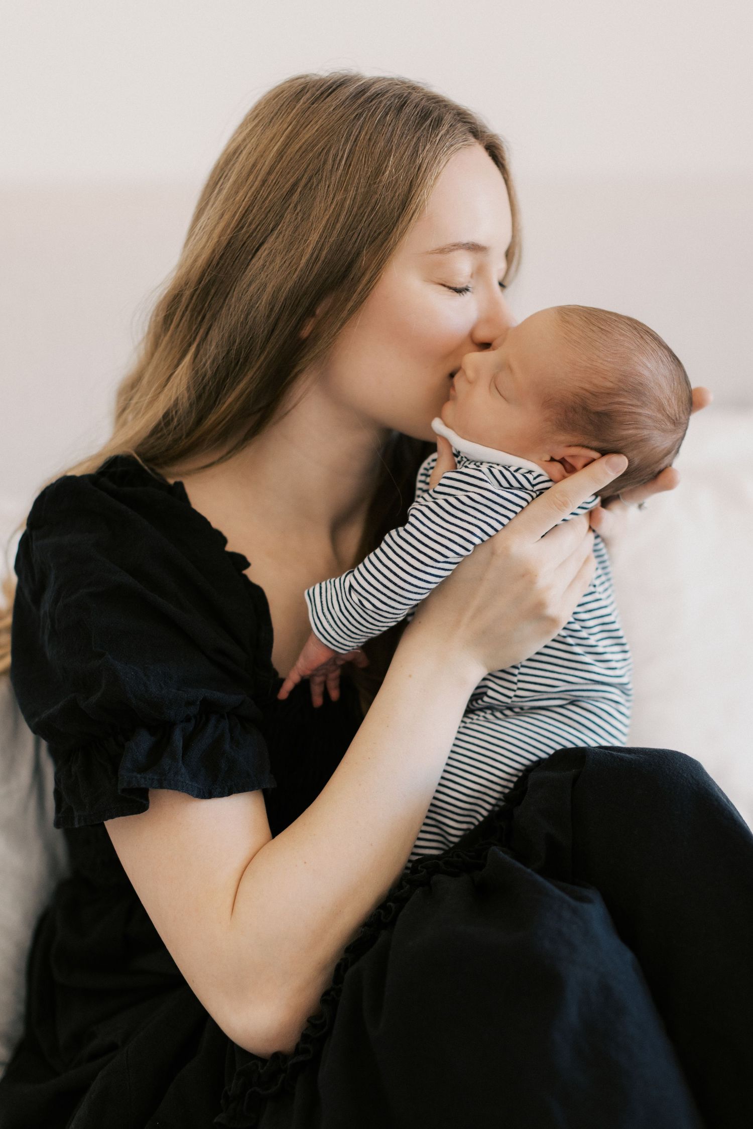 A tender mother in a black dress gently kisses her newborn baby dressed in a striped onesie.