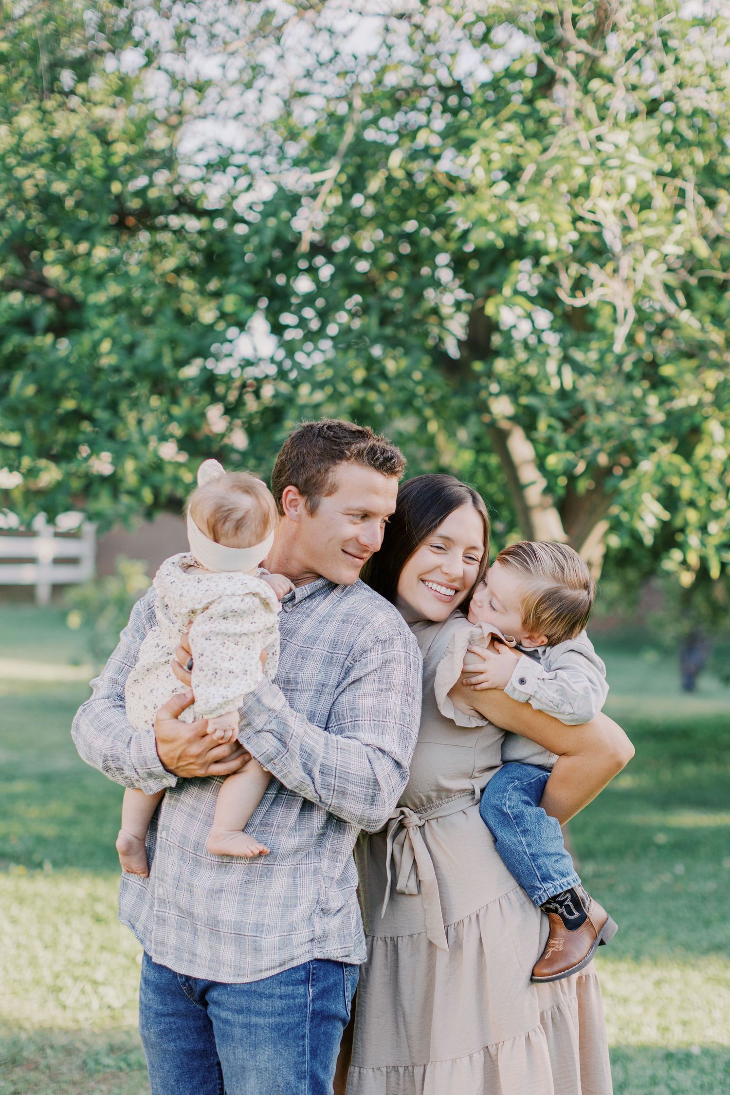 Happy family of four embracing outdoors, parents holding two young children while smiling in a lush green park setting.