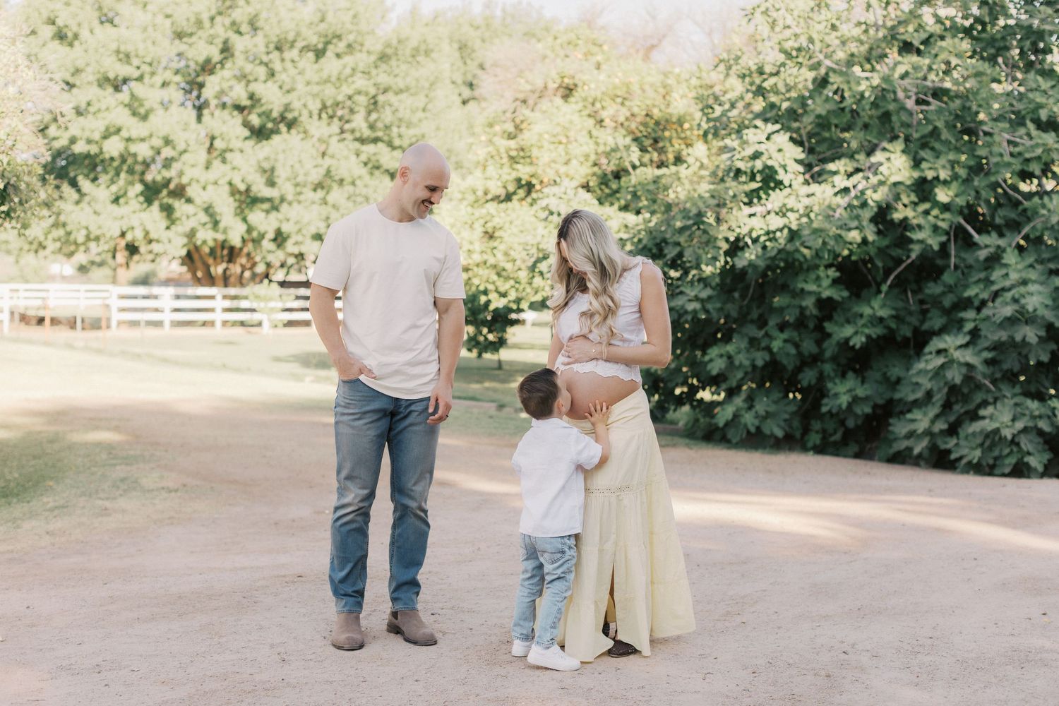 Pregnant mother in yellow dress stands with husband and toddler son in a sunny outdoor farm setting.