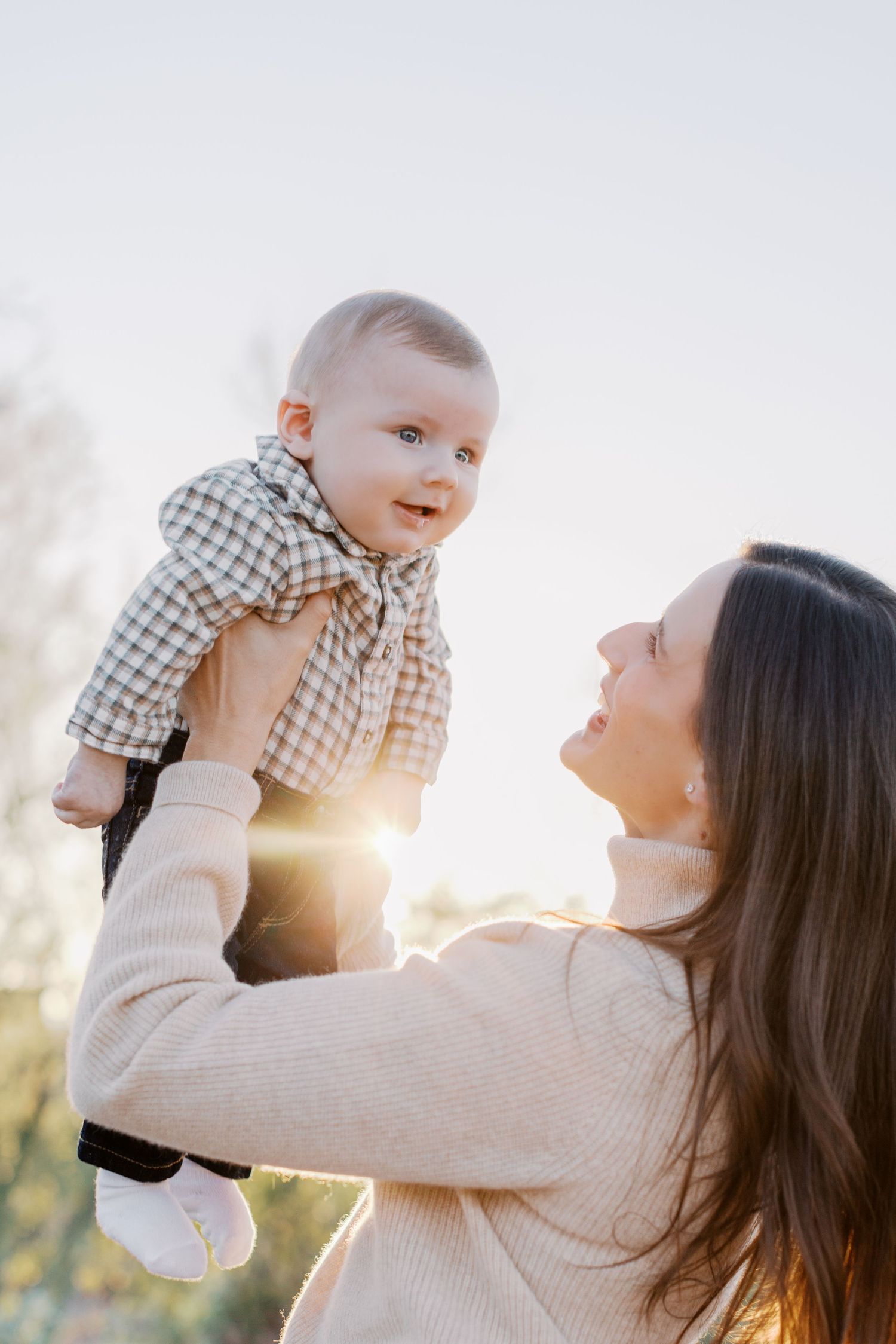 Mother lifting smiling baby boy overhead against golden sunset backlight outdoors.