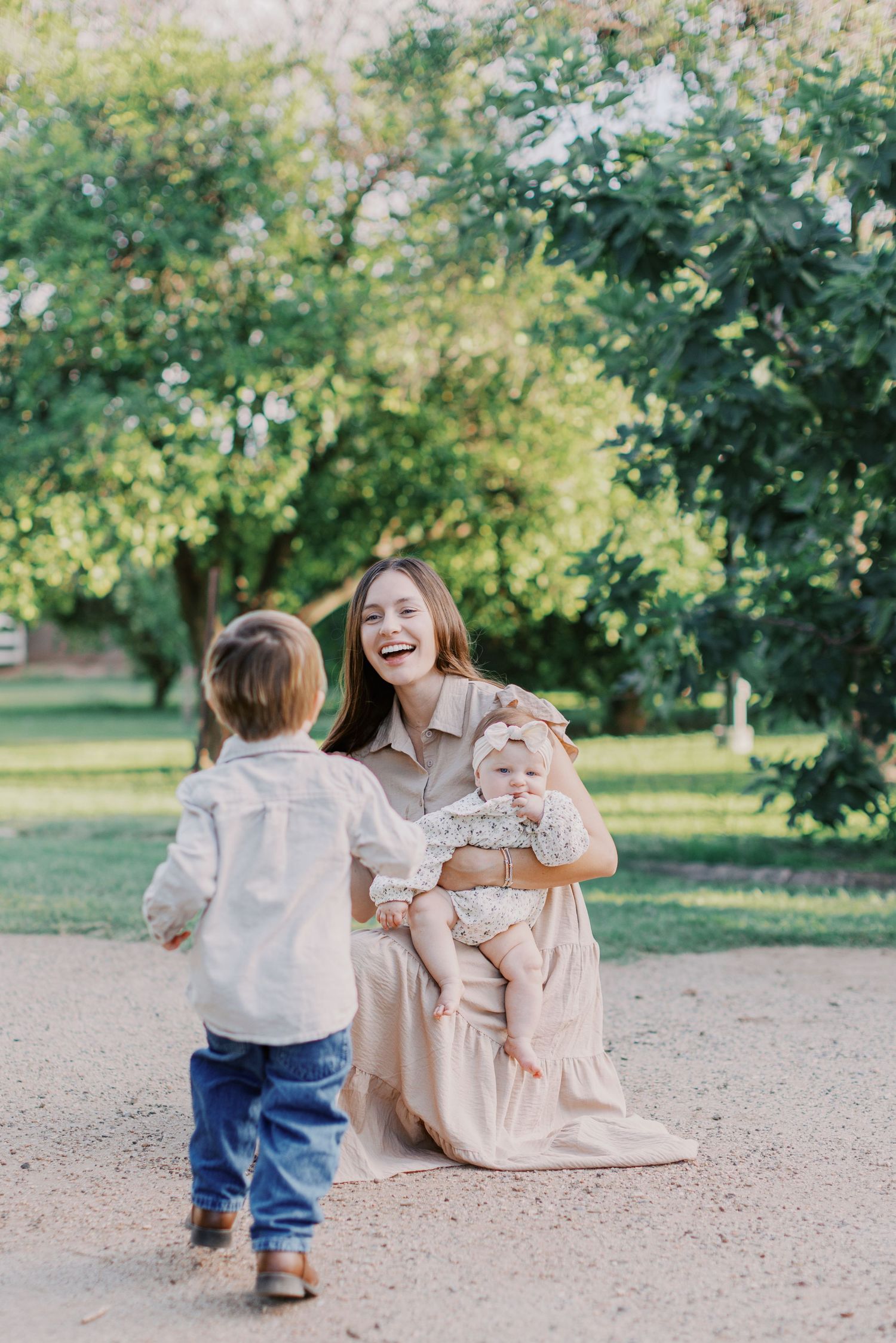 A laughing mother in a beige dress holds her baby girl while her toddler son runs toward her in a sunny park.