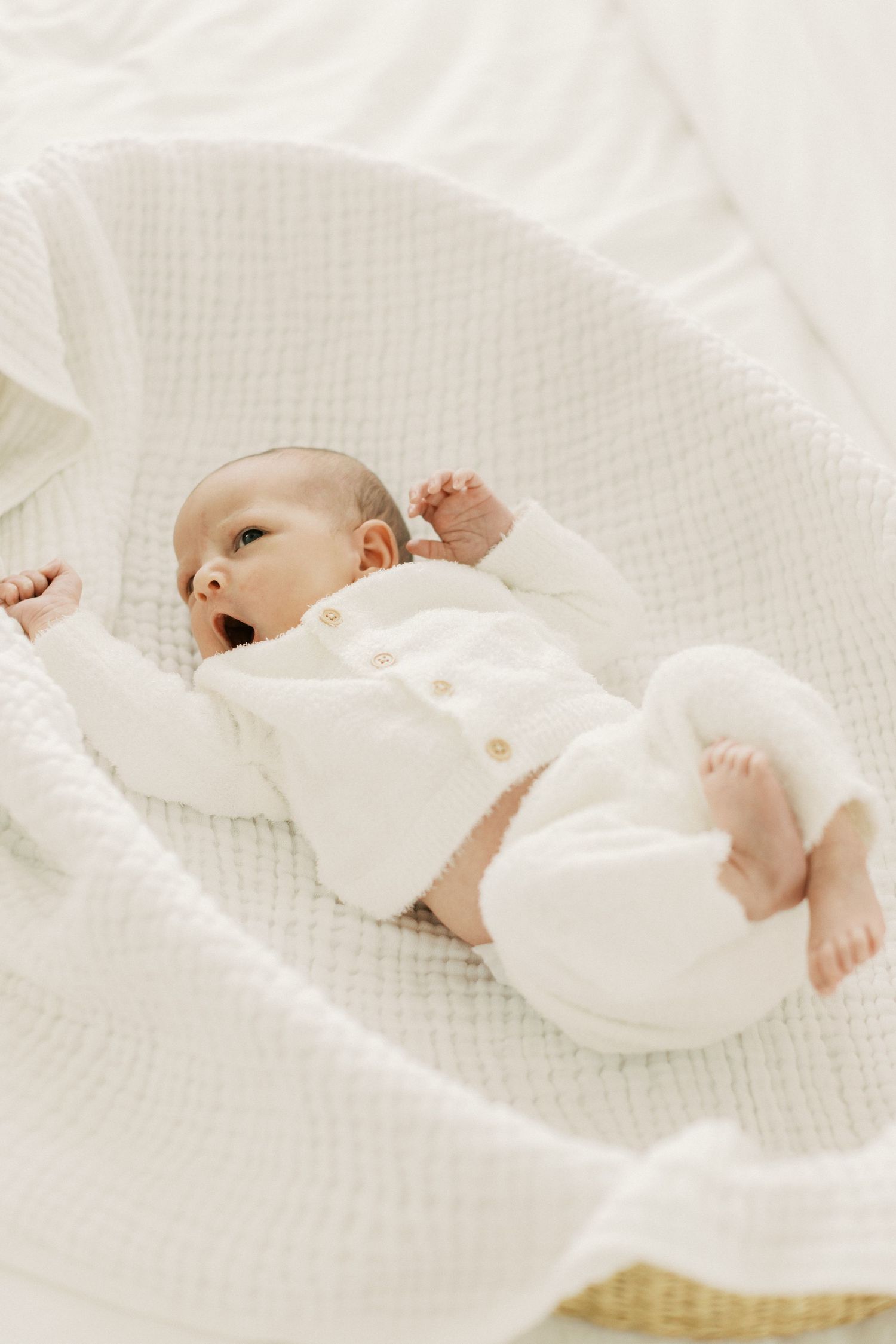 Newborn baby in white knit outfit lying in a wicker Moses basket with soft white blanket.