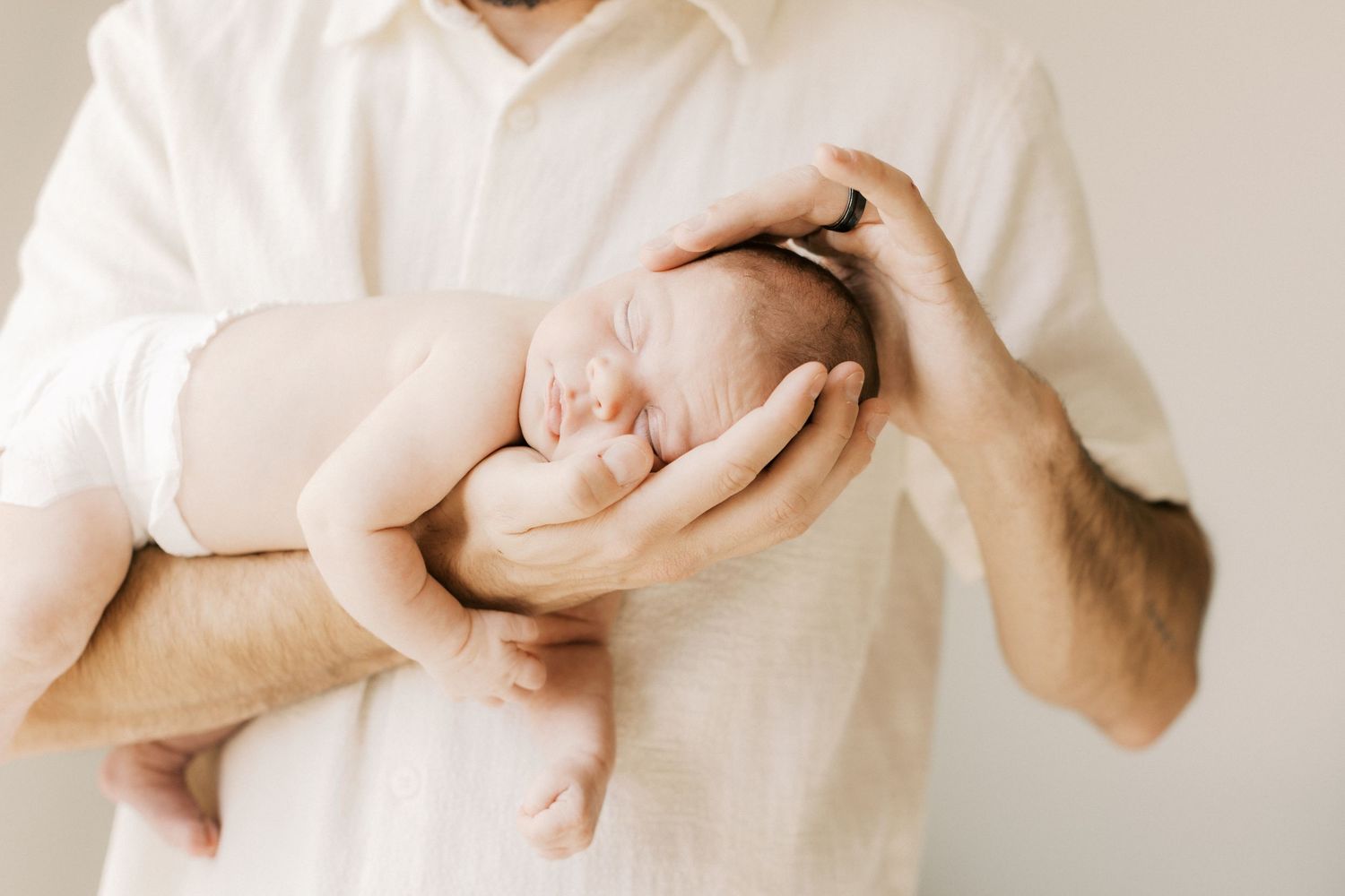 A father tenderly cradles a sleeping newborn baby in both hands against a soft white background.