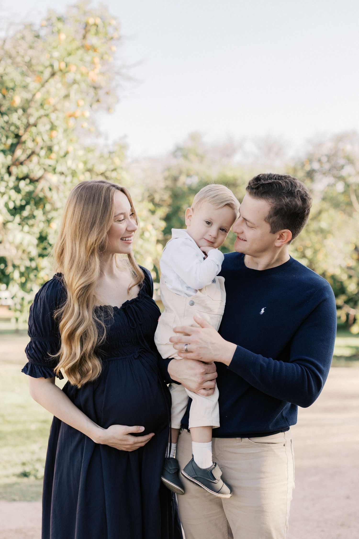 Pregnant woman in navy dress with husband holding toddler son during outdoor maternity photo session.