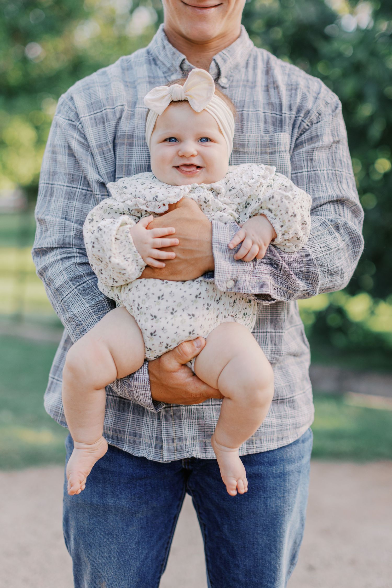 A smiling baby girl in a floral outfit and bow headband being held by a man in a plaid shirt outdoors.
