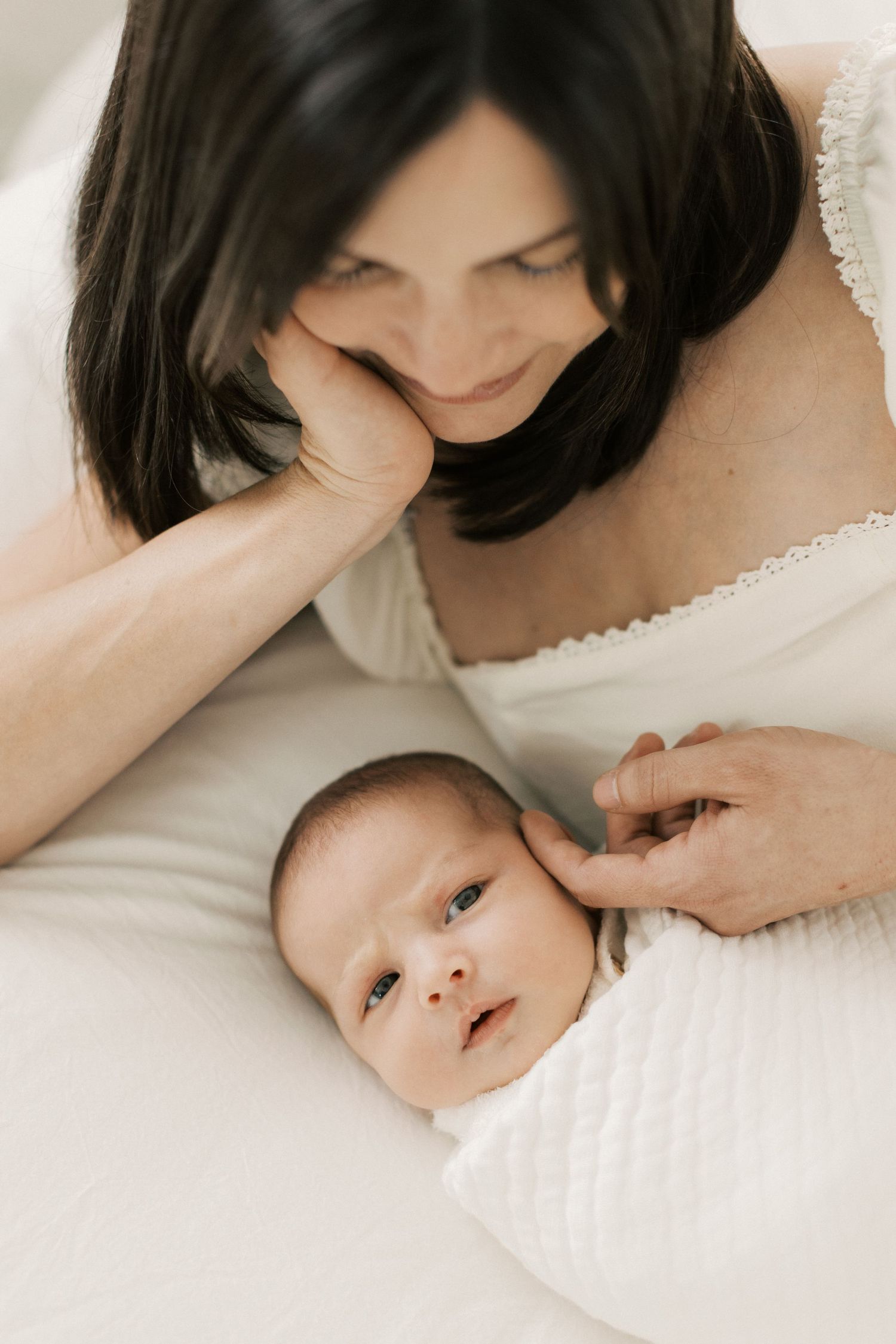 Dark-haired mother in white dress gazing lovingly at her swaddled newborn baby lying on a white bed.