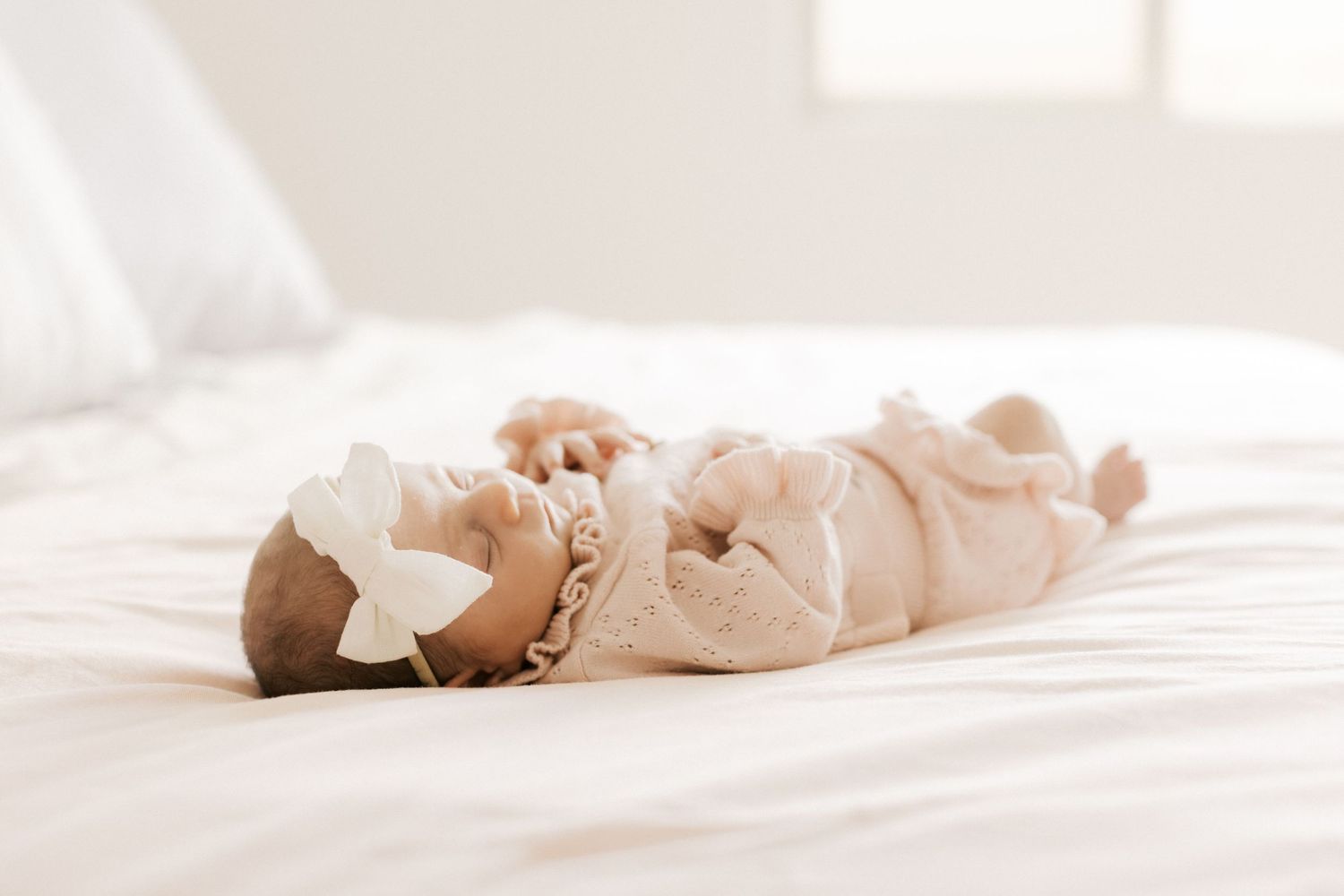 Newborn baby in white lace outfit and bow headband lying on white bed.