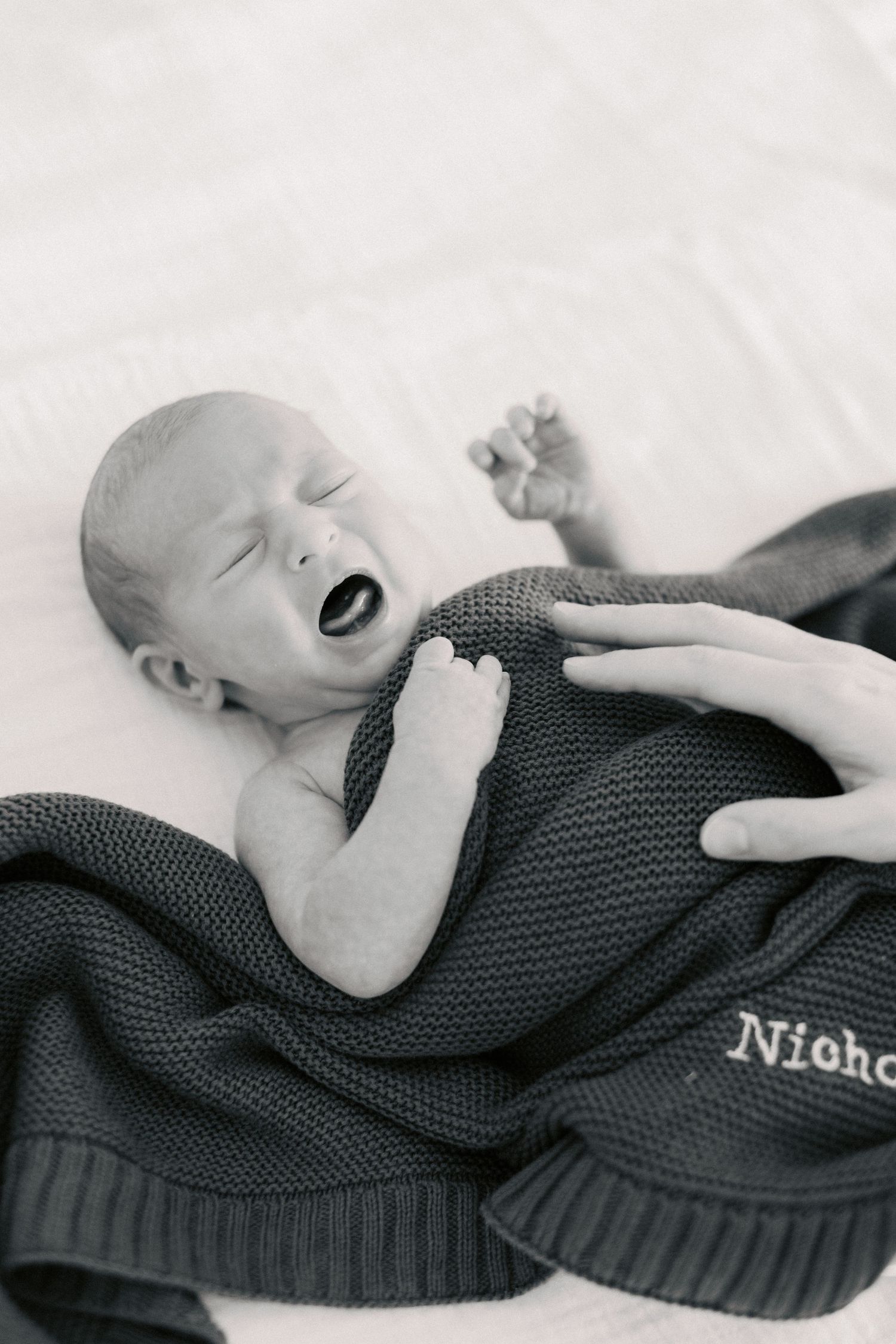 Black and white photo of newborn baby yawning wrapped in knit blanket embroidered with name Nicho.