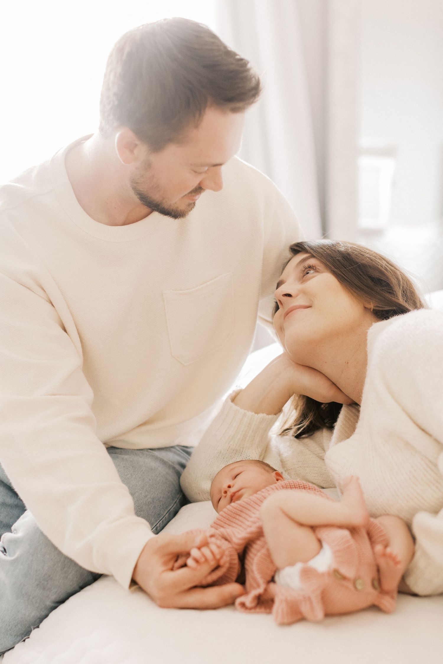 A couple in white sweaters lovingly gazes at each other while their newborn baby rests peacefully between them.