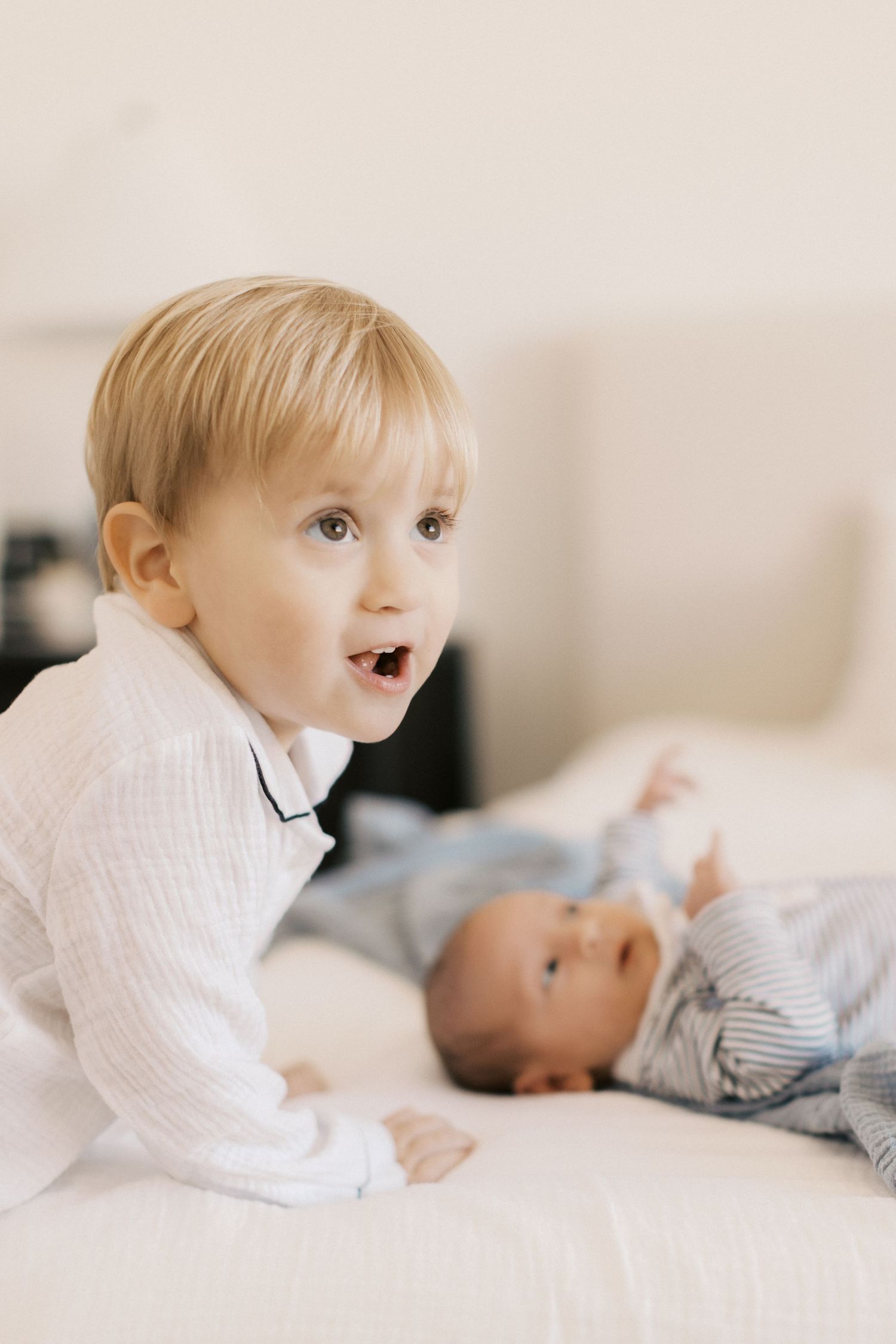 Excited toddler boy in white pajamas on bed with newborn baby sibling in blue striped outfit.