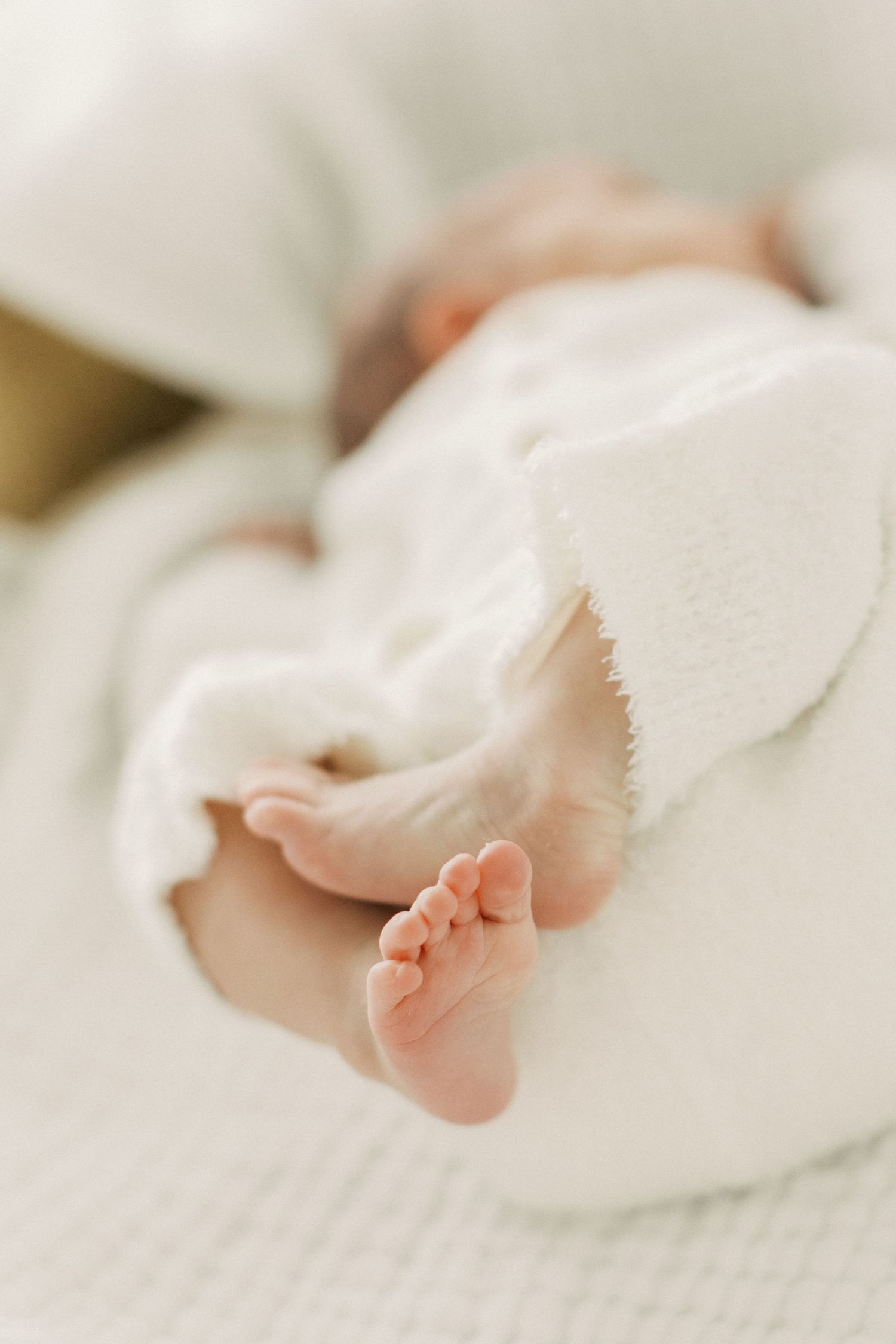 Close-up of tiny newborn baby feet peeking out from a soft white blanket on a white surface.