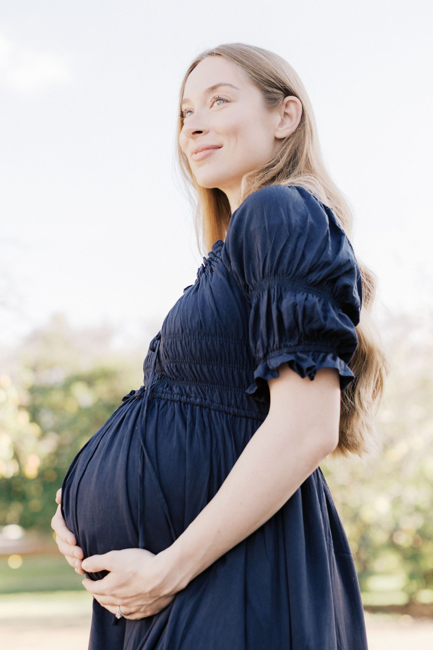 Smiling pregnant woman in navy blue dress cradling her baby bump outdoors in a sunlit park.