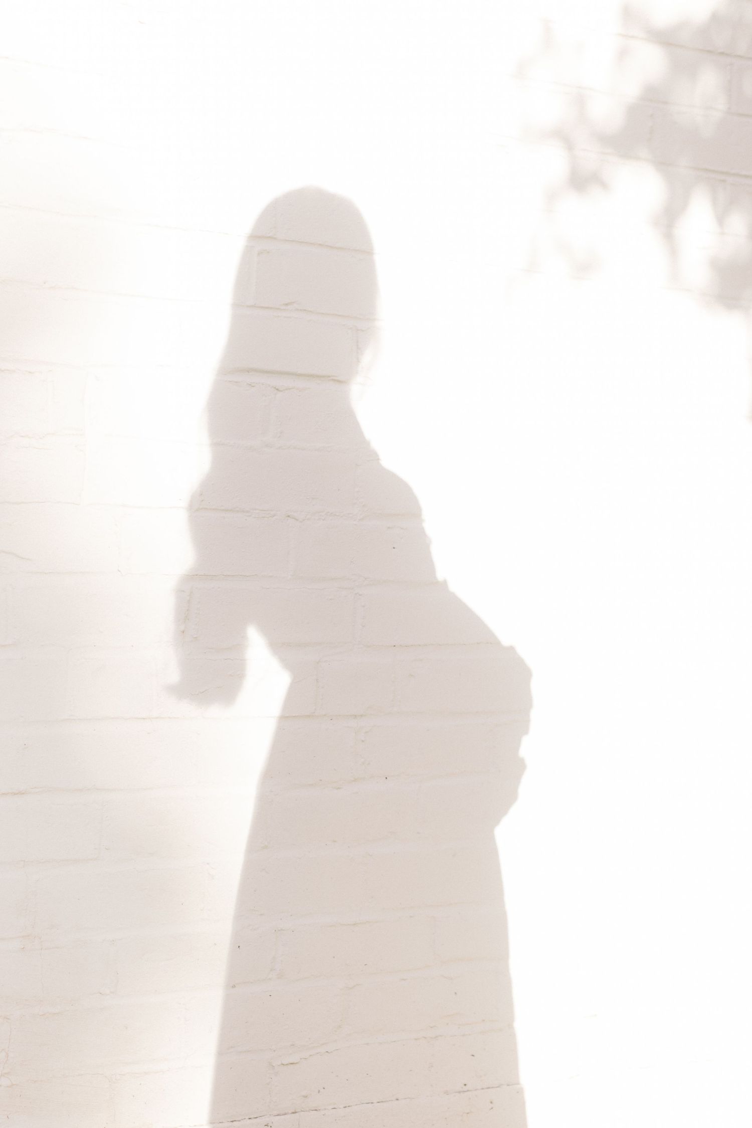 Shadow of a pregnant woman silhouetted against a bright white brick wall in soft natural light.