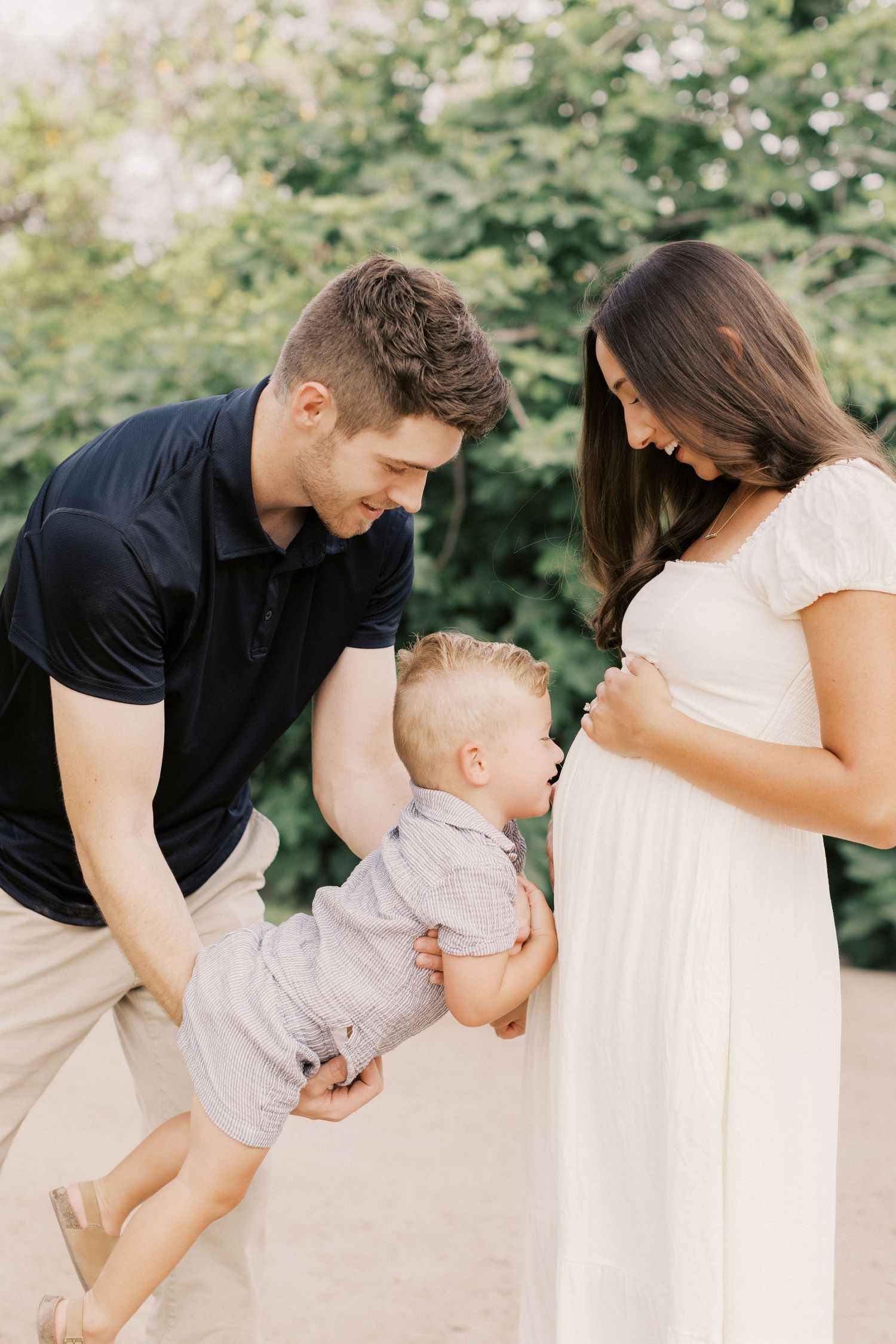 Pregnant woman in white dress with husband and toddler son touching her baby bump outdoors.