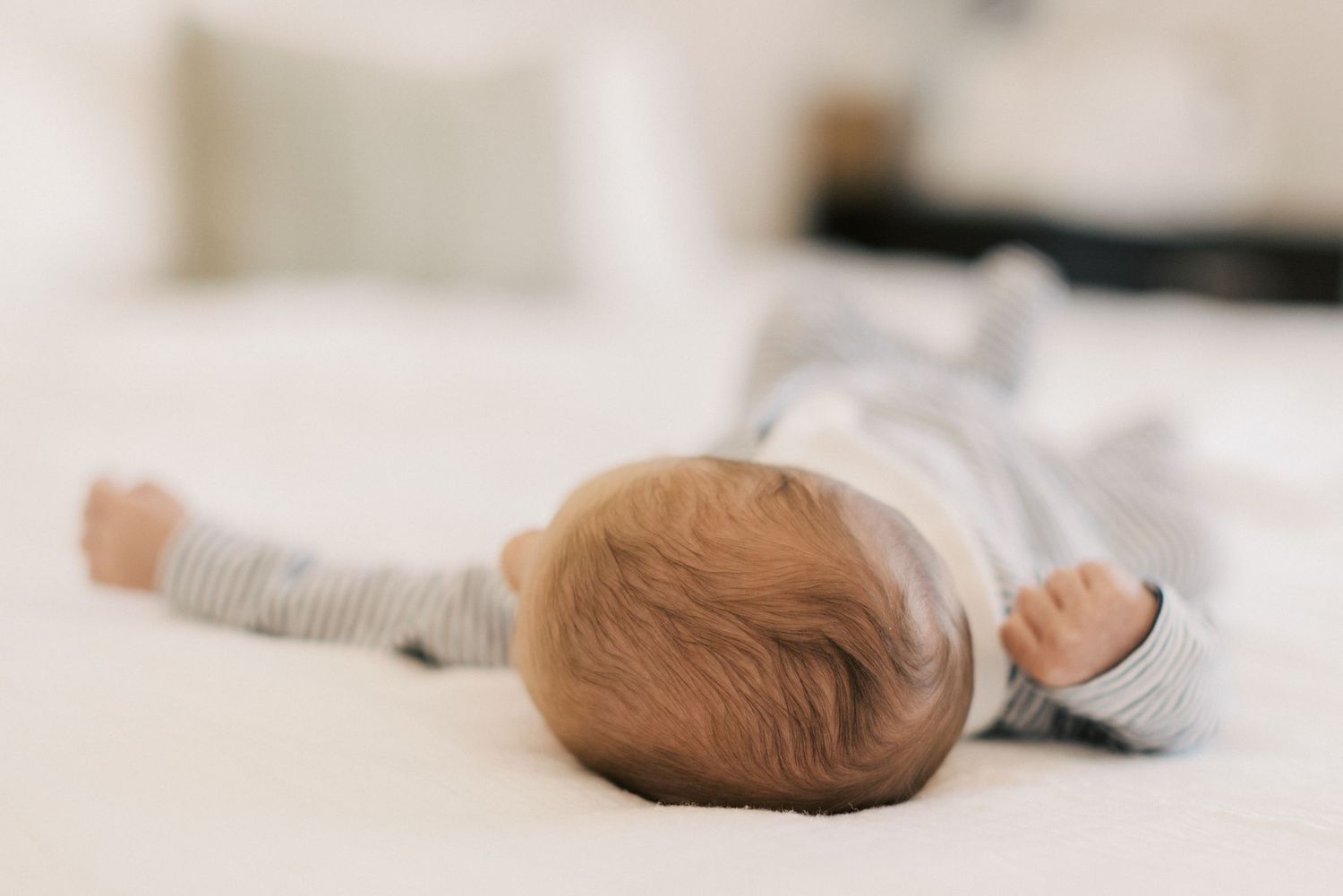 Newborn baby in gray striped onesie lies face down on a white bed with arms stretched out.