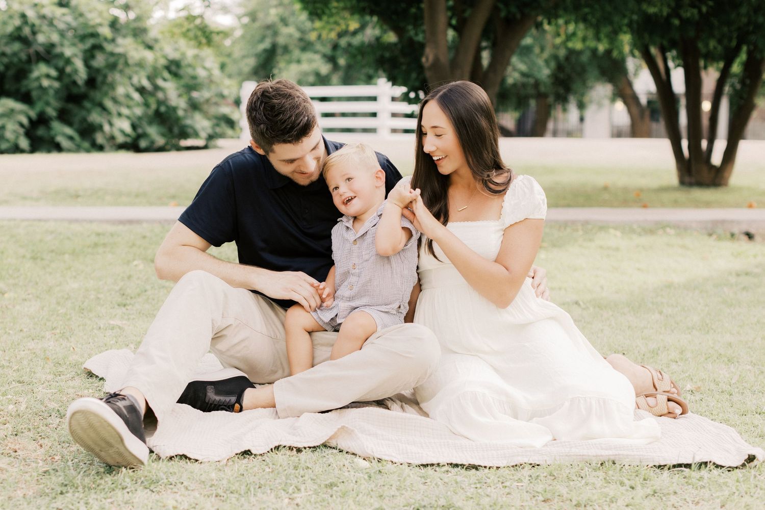Family of three sitting on blanket in park with toddler wearing birthday hat outdoors.