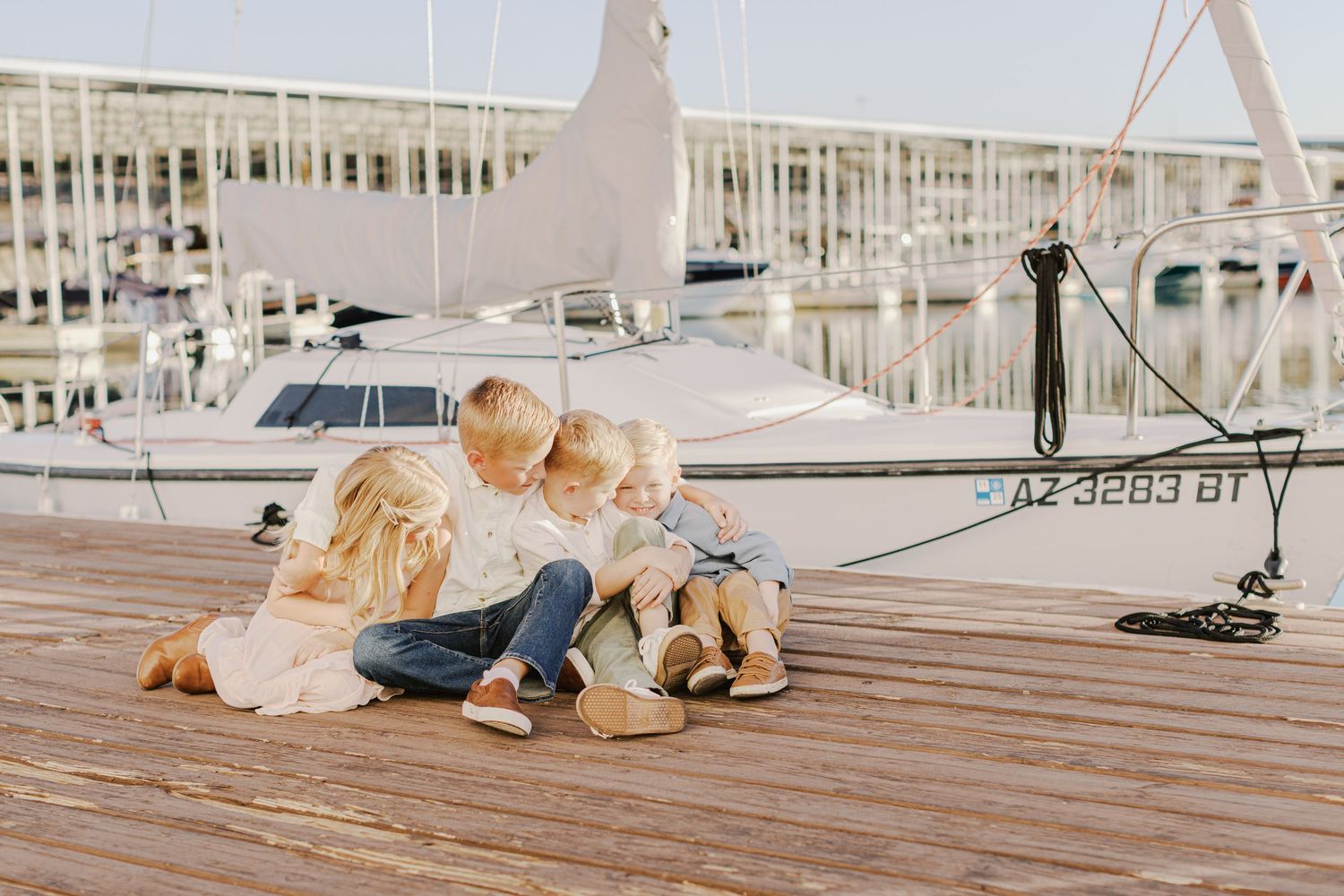 Three young siblings sitting together on a sunny marina dock beside a sailboat.
