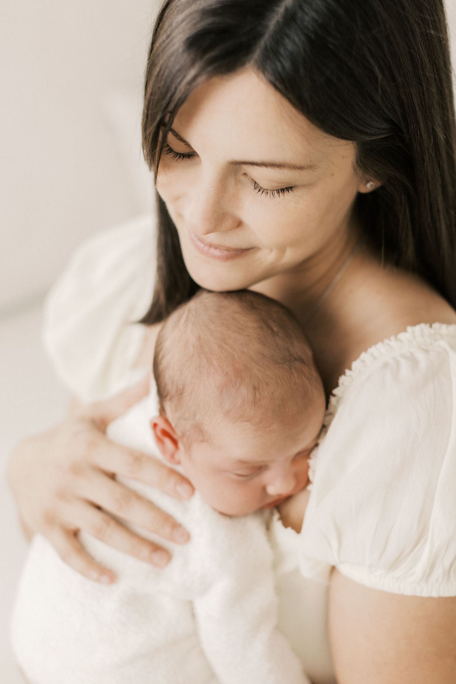 Gentle maternal moment with newborn baby in white against light background.