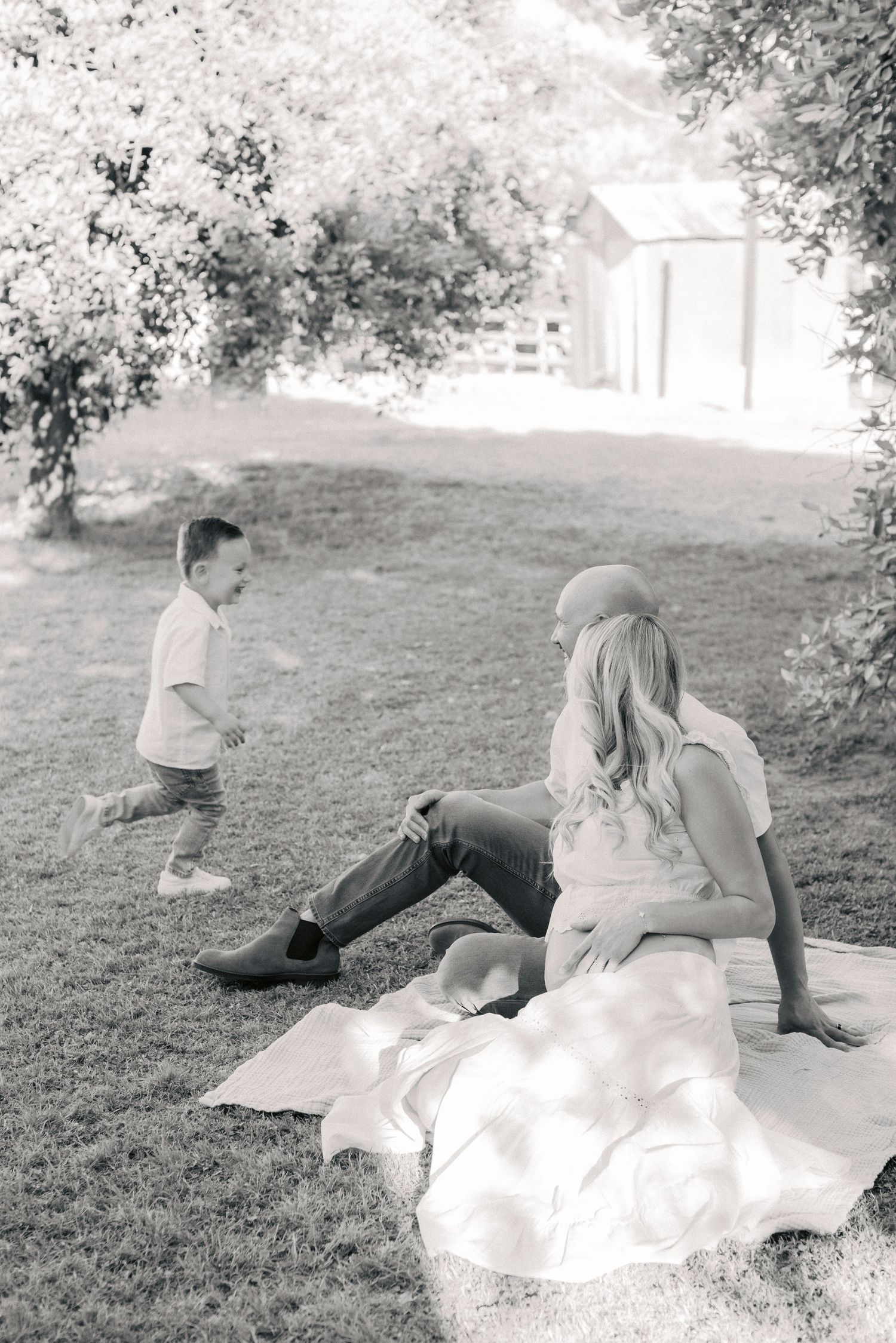 Black and white photo of young mother relaxing on blanket in yard while energetic toddler son runs nearby.