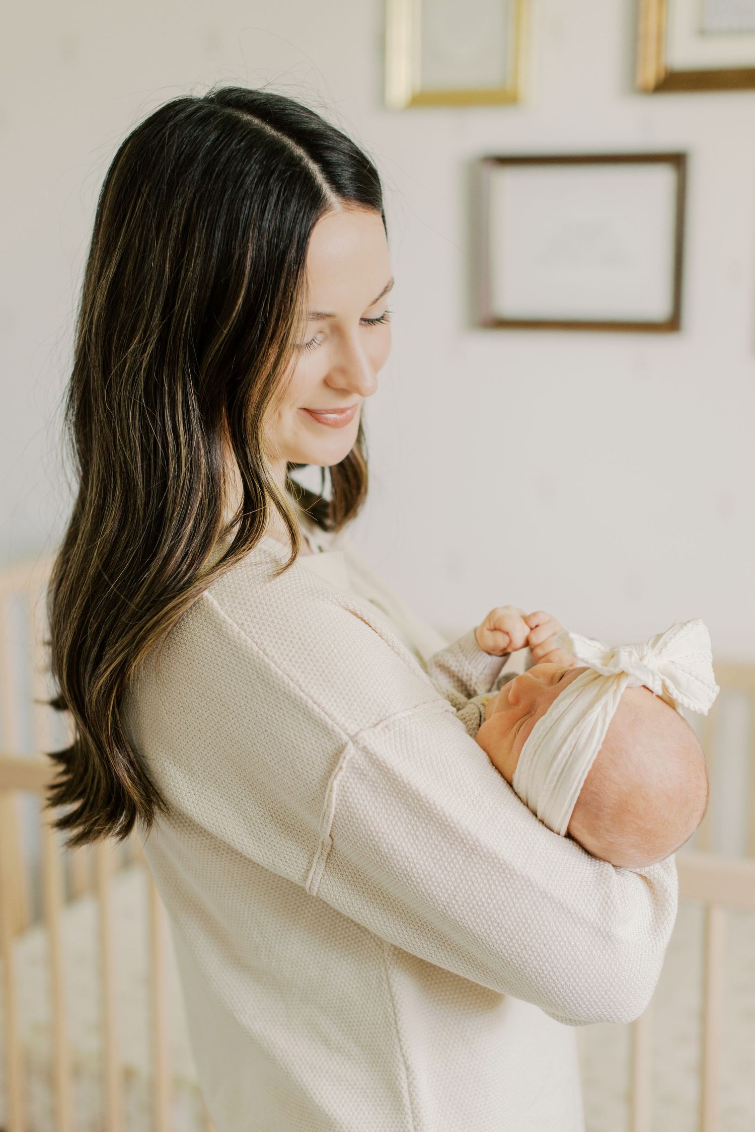 A dark-haired mother in a cream sweater tenderly holds her sleeping newborn baby in a softly lit nursery.